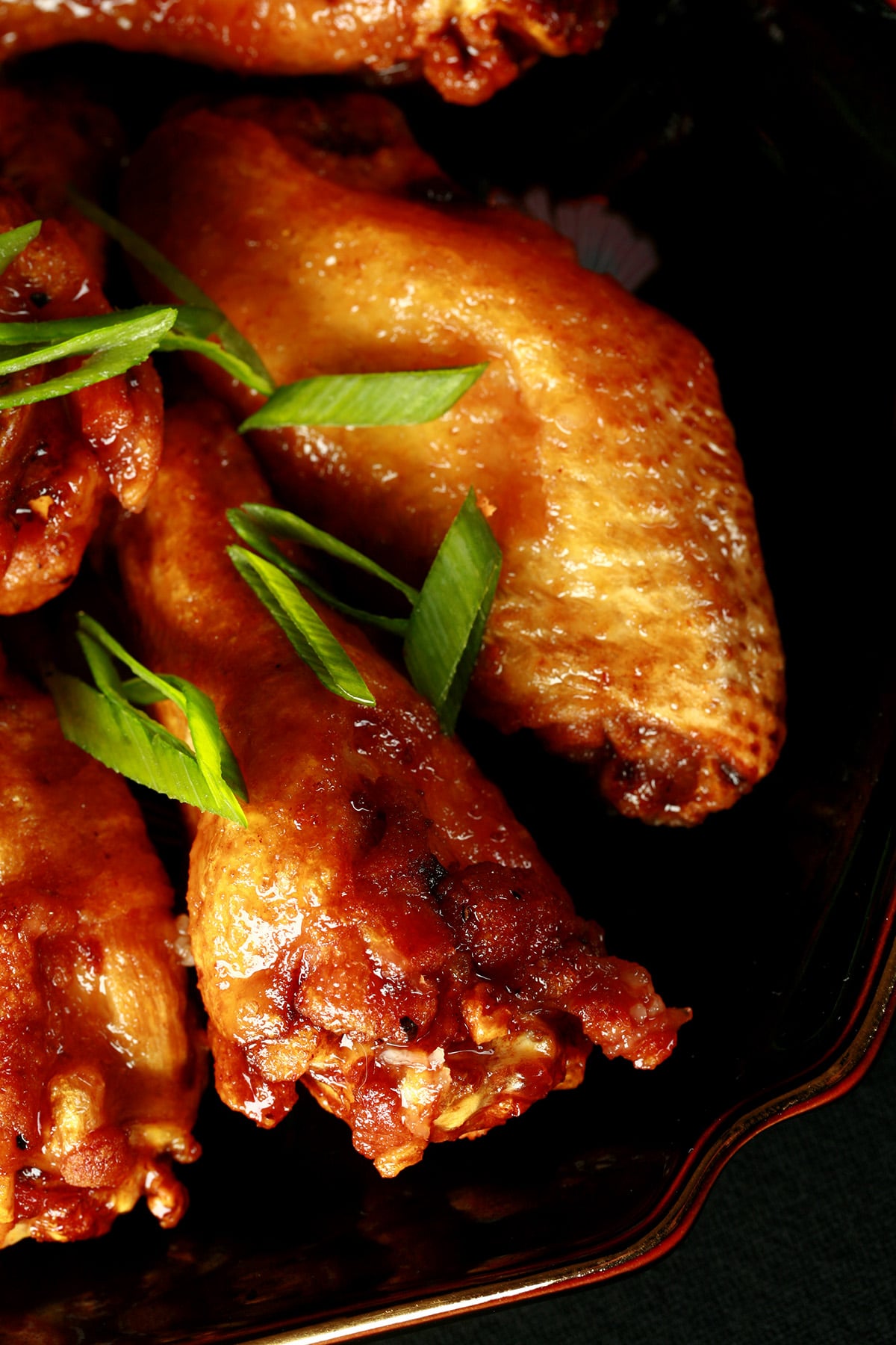 A small black plate with a pile of Spicy Ginger Glazed Wings piled on it, topped with slices of green onion.