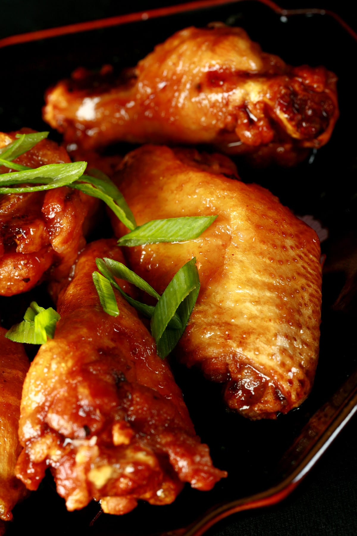 A small black plate with a pile of Spicy Ginger Glazed Wings piled on it, topped with slices of green onion.