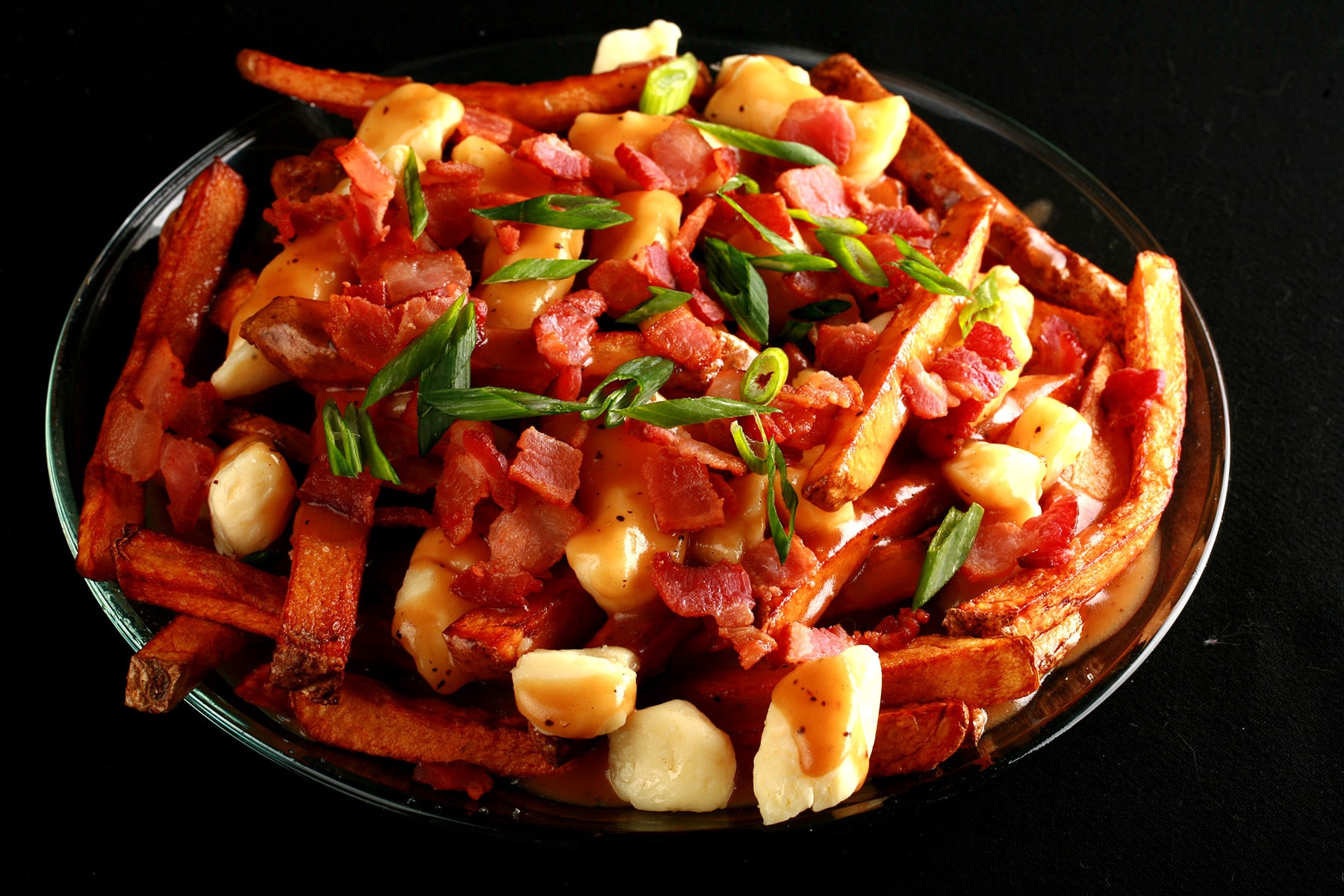 A close up photo of a plate of poutine, garnished with bacon and green onions..