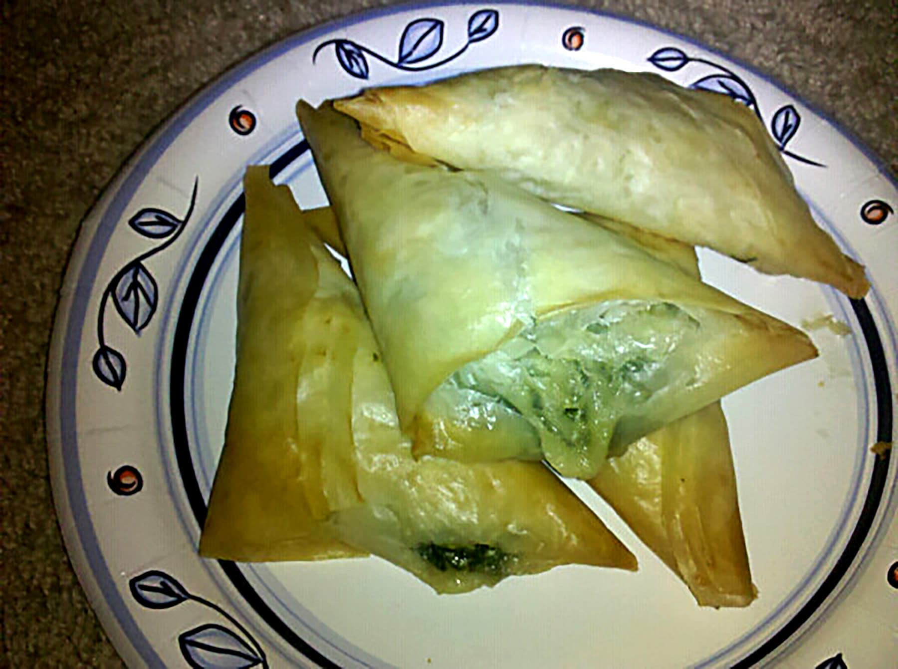 4 spanakopita pastries on a white paper plate with a blue floral border.