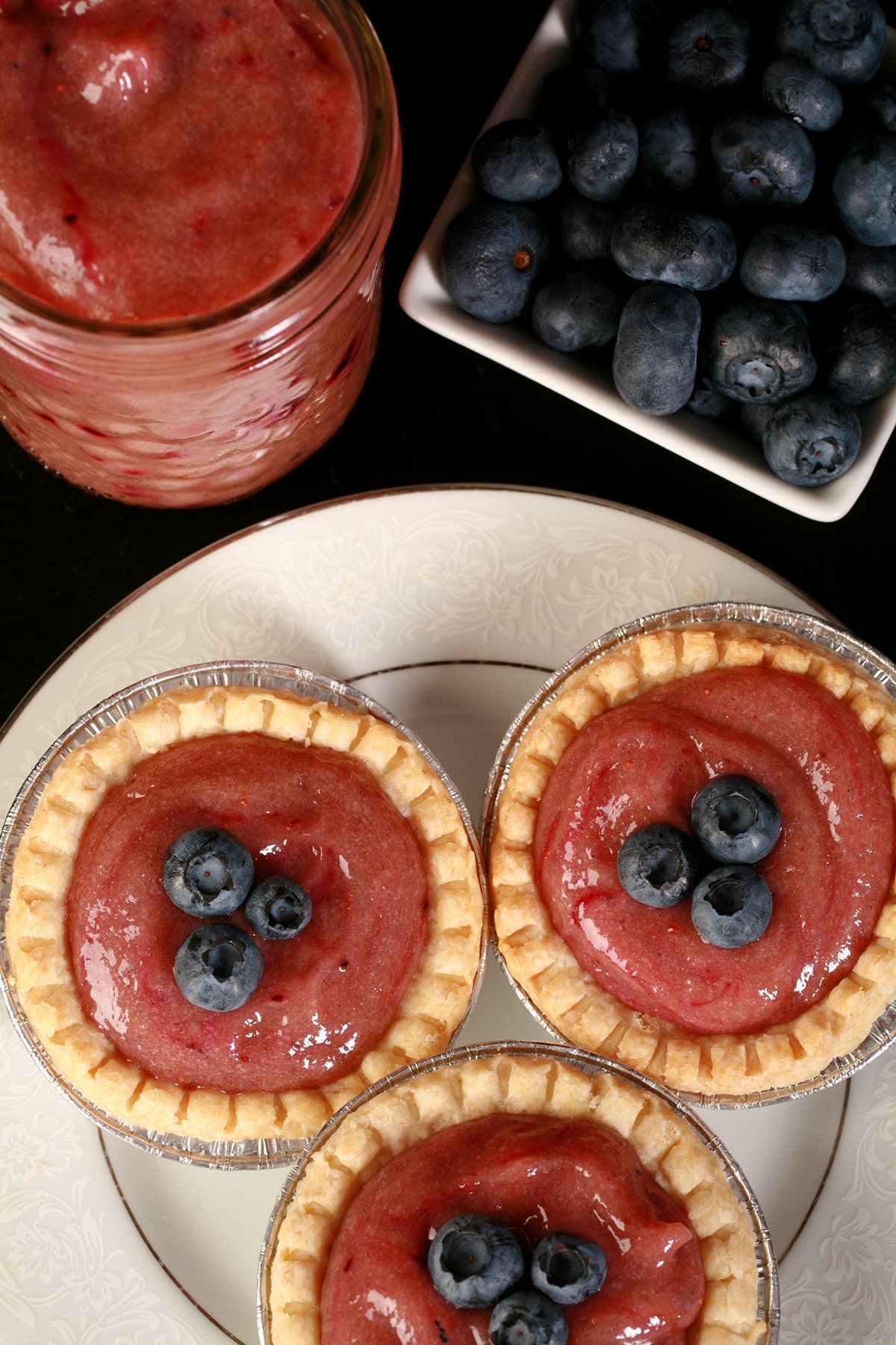 3 blueberry tarts on a plate, next to a jar of curd and a bowl of blueberries.