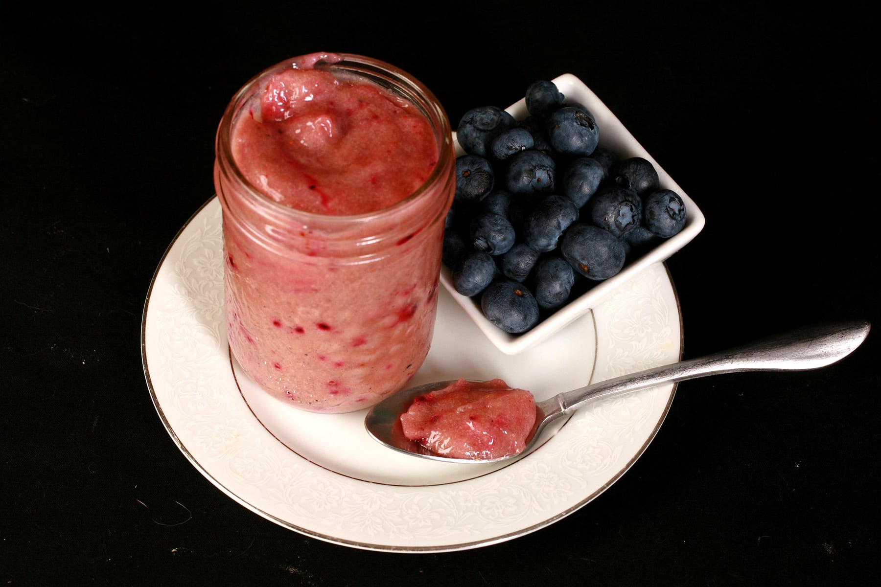 A jar of blueberry curd and a spoon of curd on a plate, along with a small bowl of fresh blueberries.