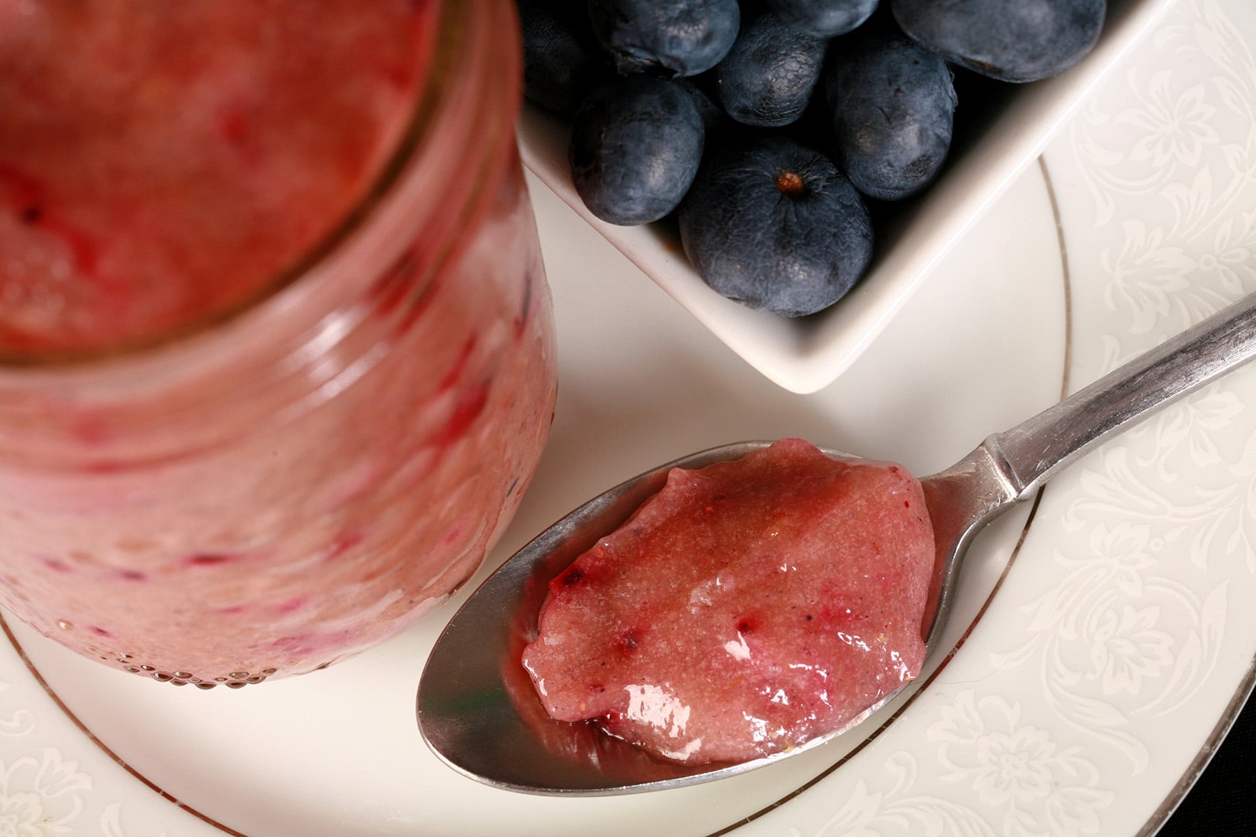 A jar of blueberry curd and a spoon of curd on a plate, along with a small bowl of fresh blueberries.