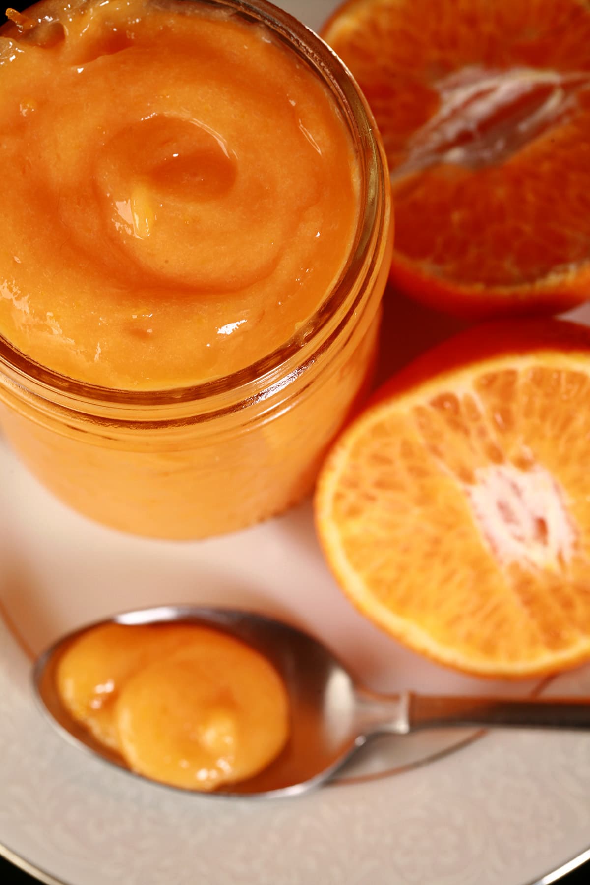 A jar of orange curd on a plate, along with a spoon of curd and a sliced orange.