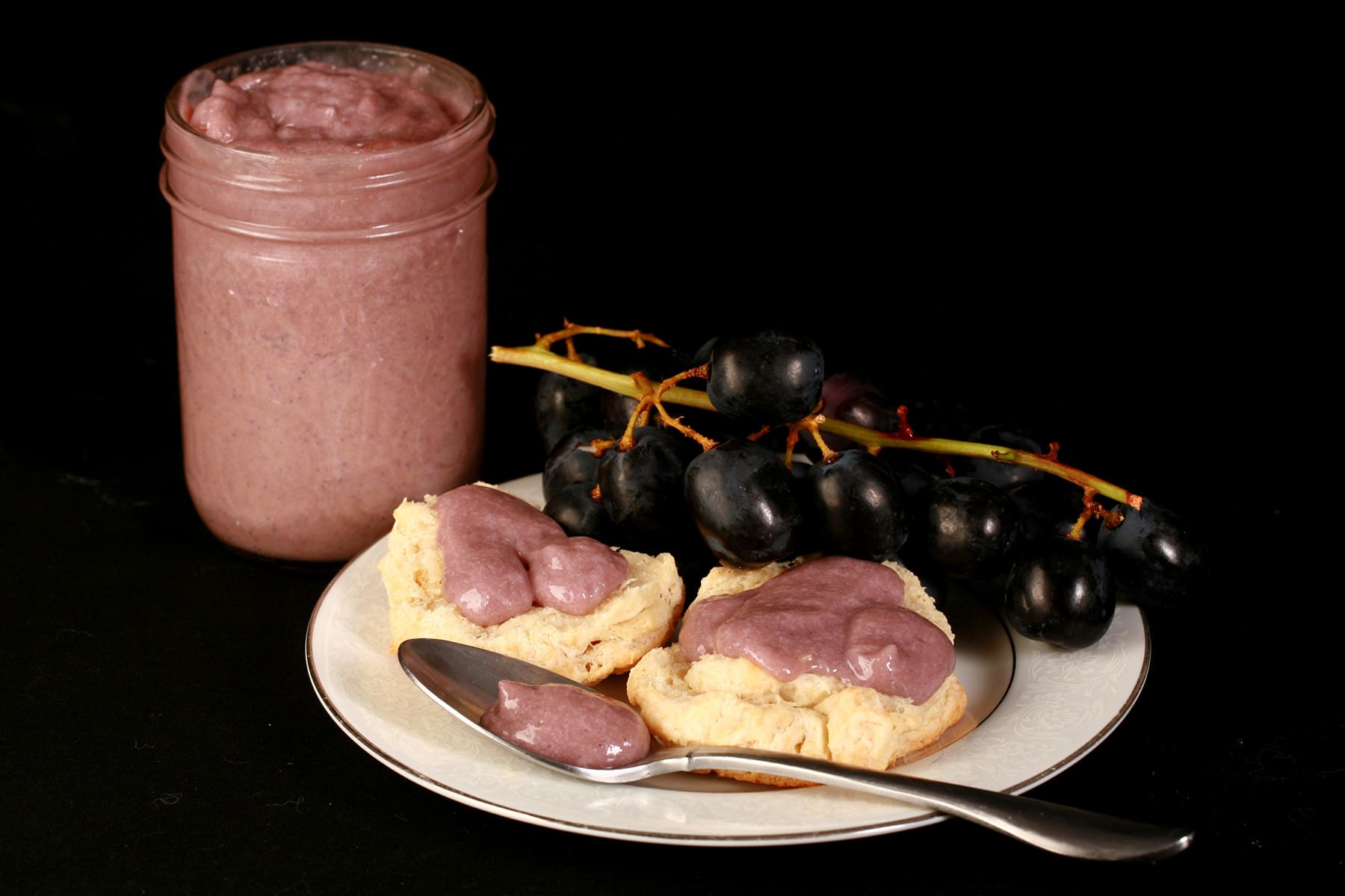 Two biscuits with grape curd on a plate, along with some concord grapes and a spoon of curd.