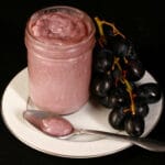 A jar of concord grape curd on a plate, along with some grapes and a spoonful of the curd.