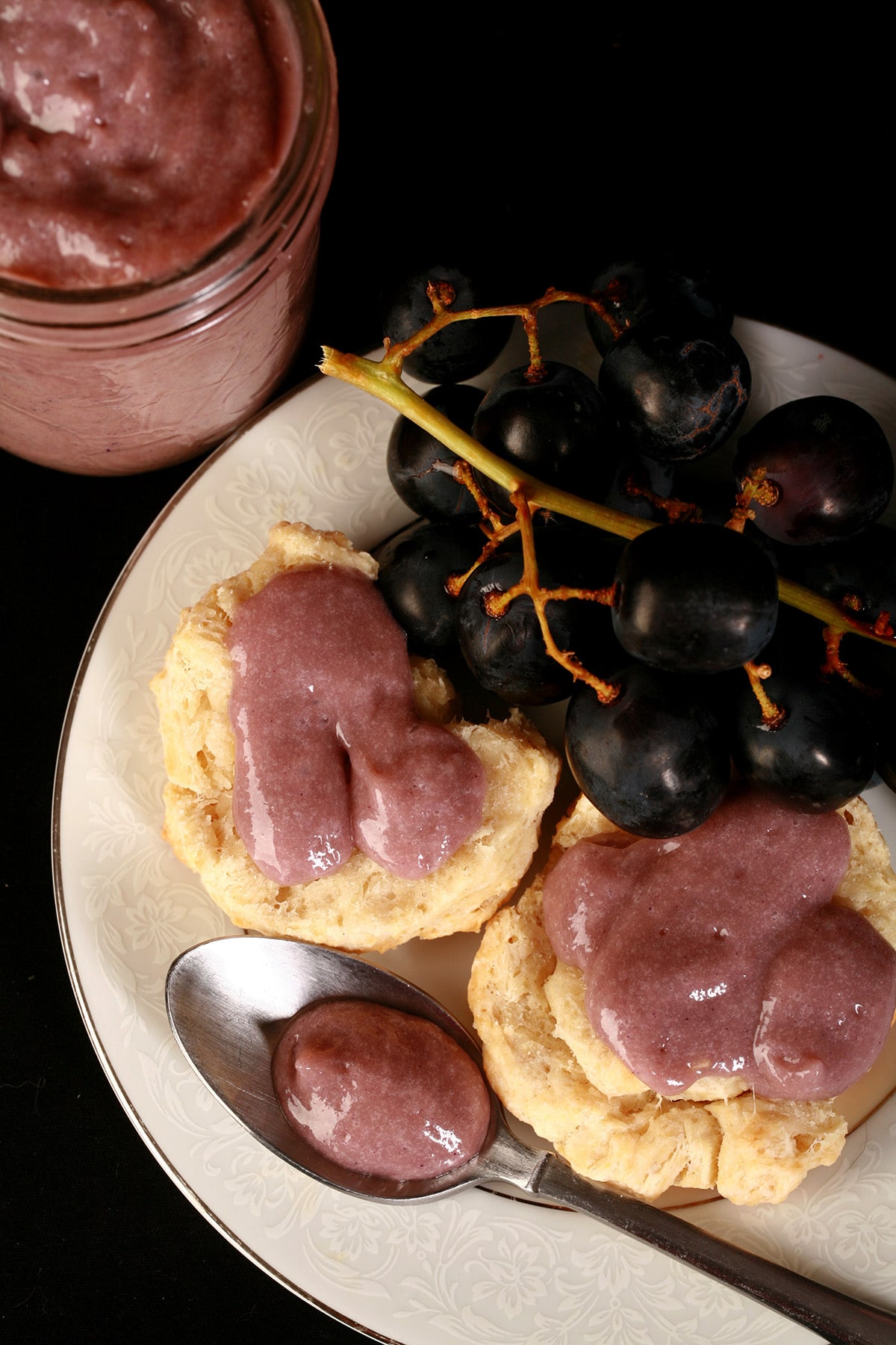 Two biscuits with grape curd on a plate, along with some concord grapes and a spoon of curd.