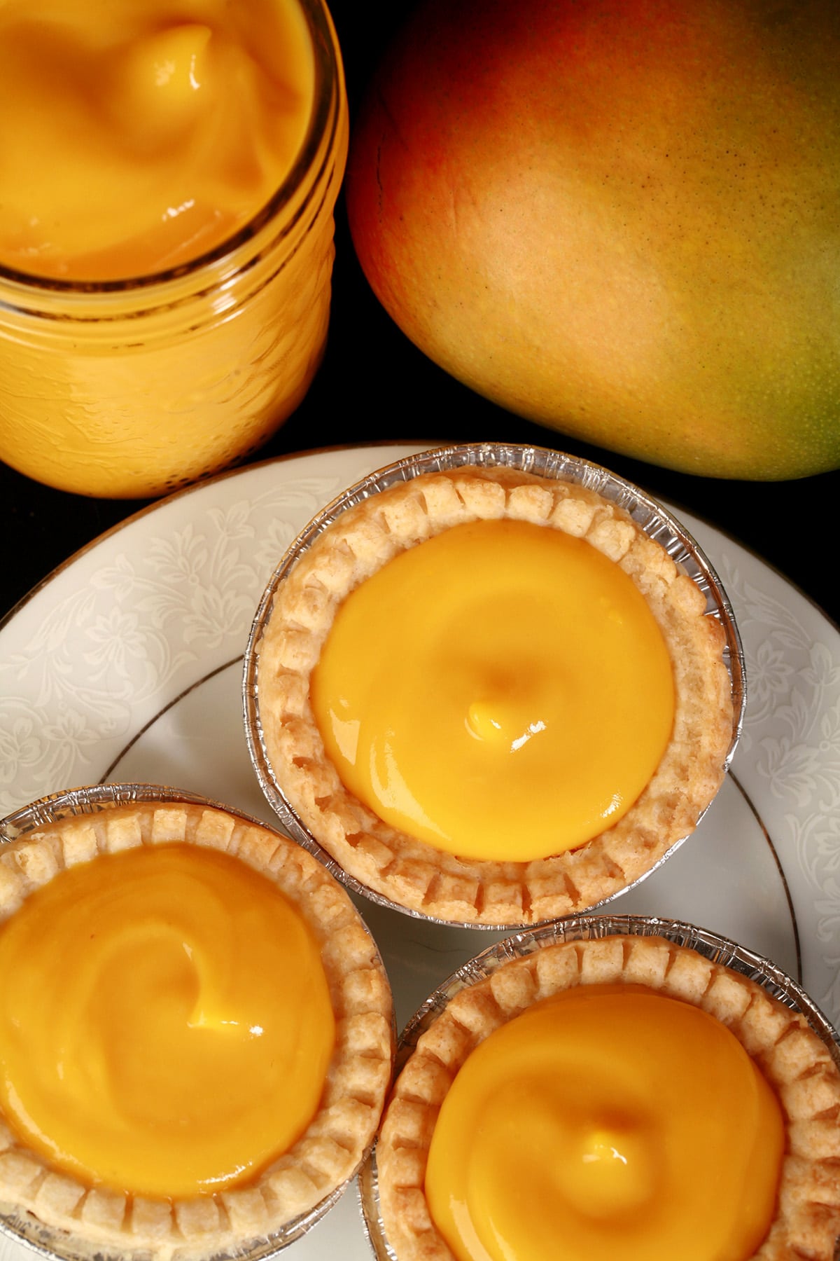 3 mango tarts on a plate, with a jar of mango curd and a mango behind the plate.