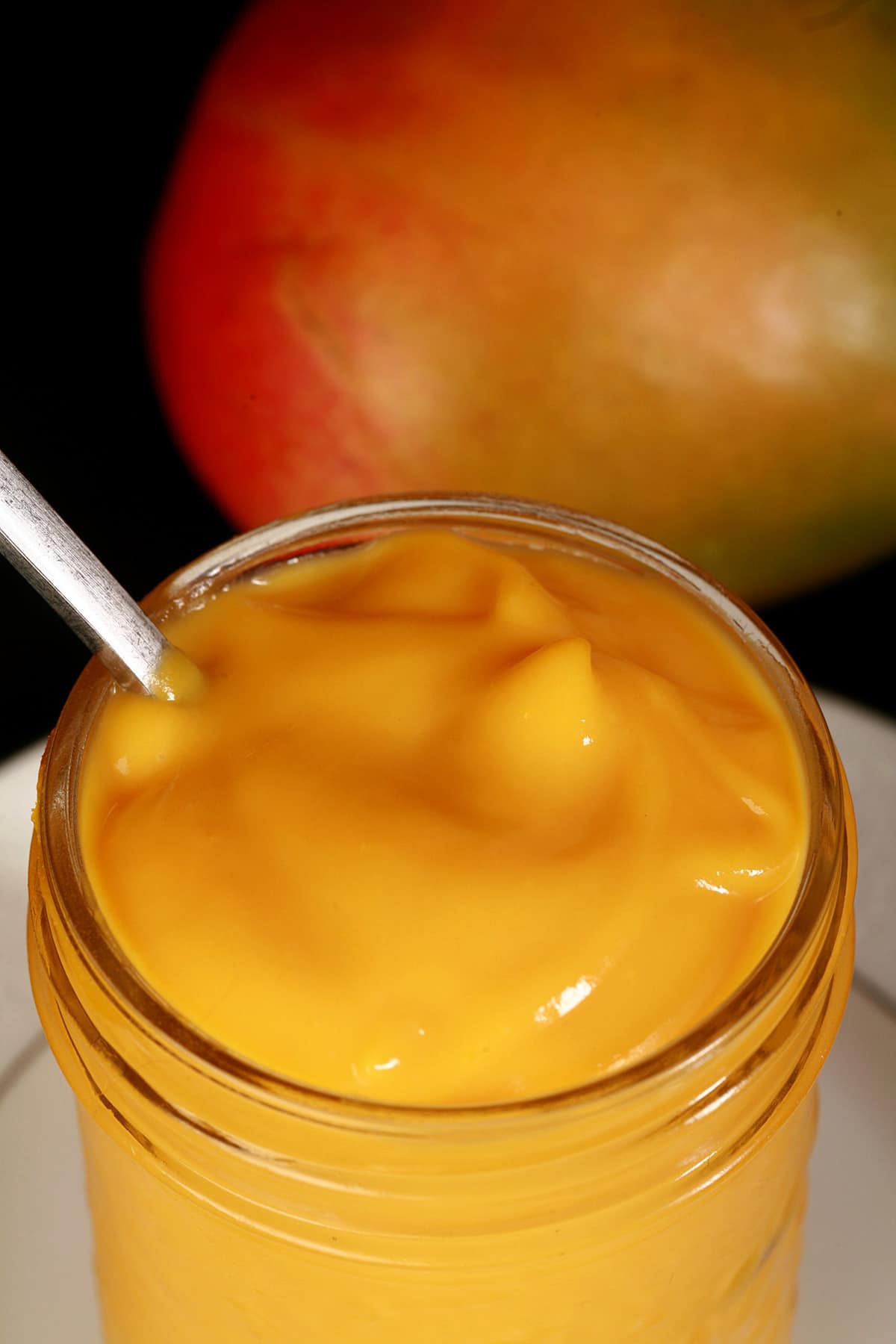 A jar of mango curd on a plate, with a whole mango behind it.