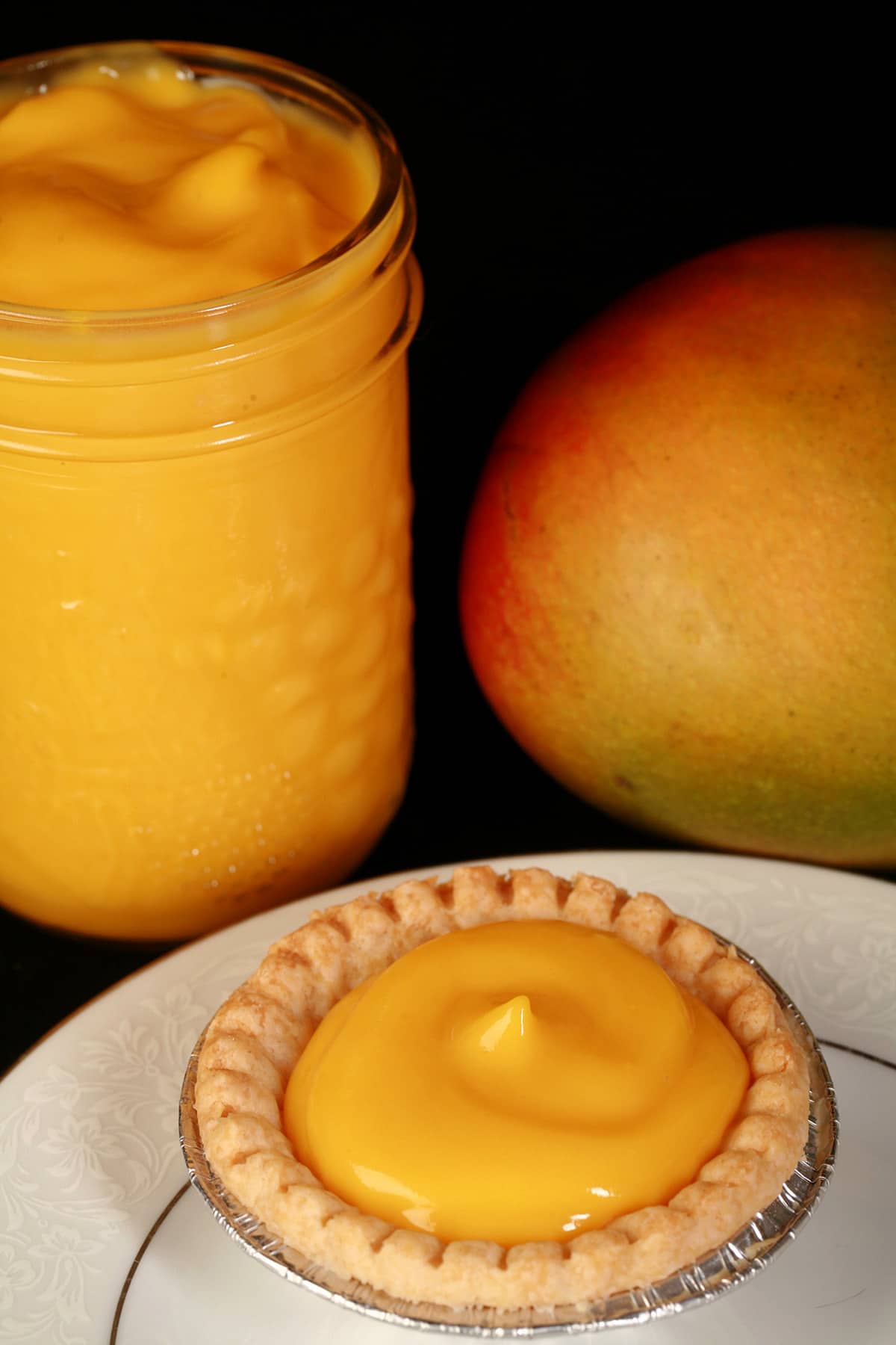 A mango tart on a plate, with a jar of mango curd and a mango behind the plate.