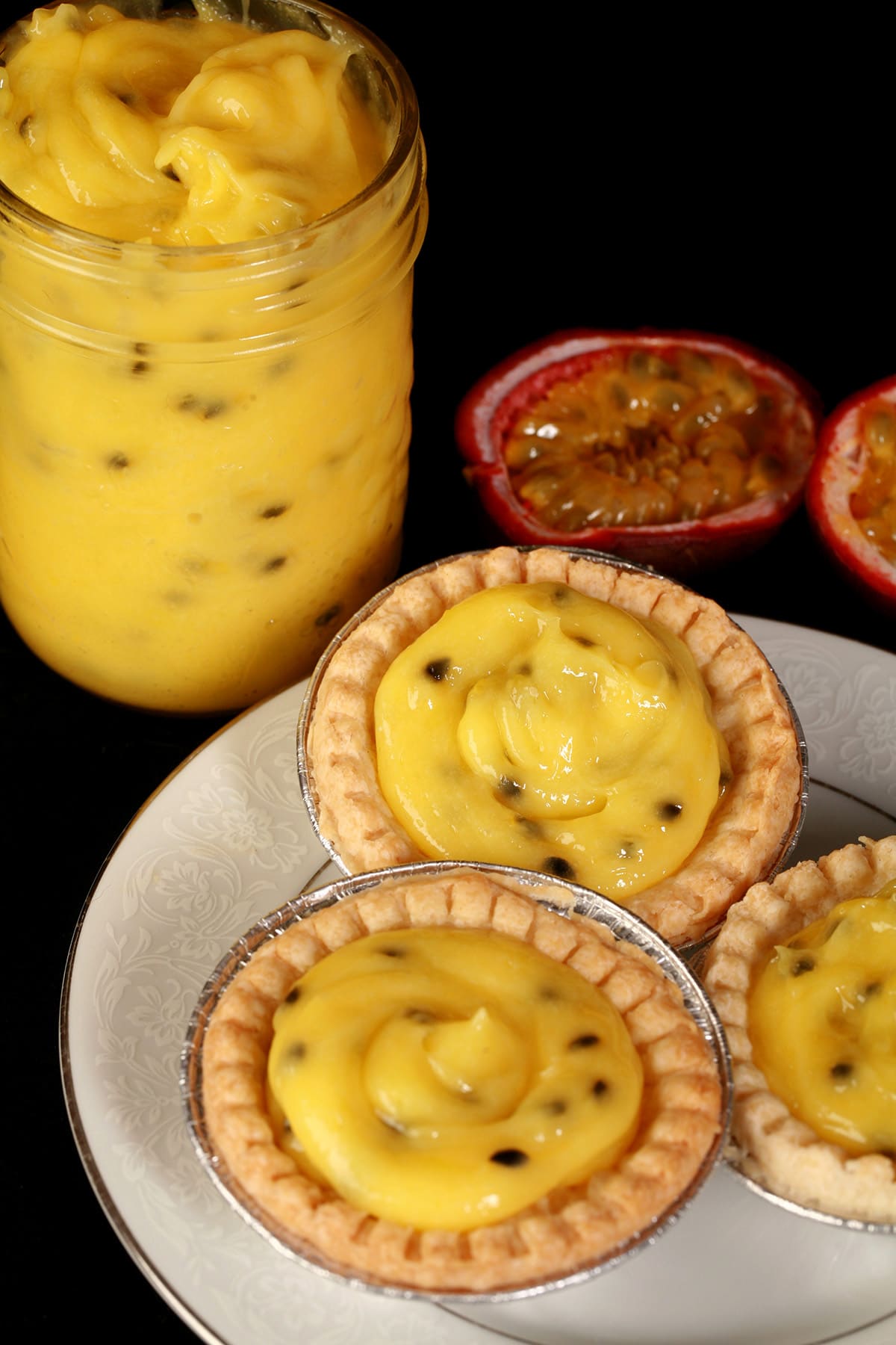 3 passionfruit tarts on a plate. There is a jar of passionfruit curd next to the plate, along with a halved passionfruit.