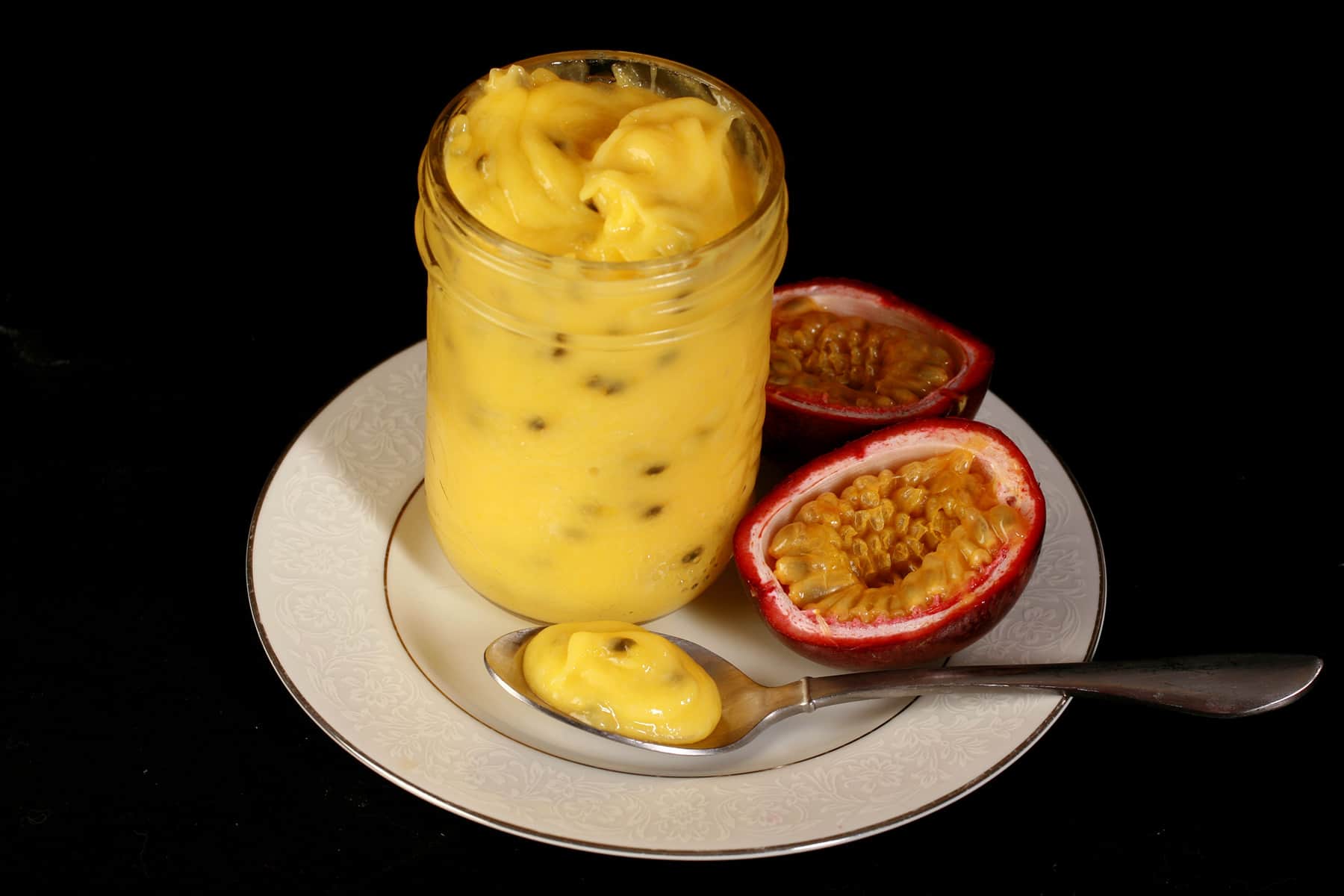 A jar of passionfruit curd on a plate, along with a spoon of curd and a sliced passionfruit.