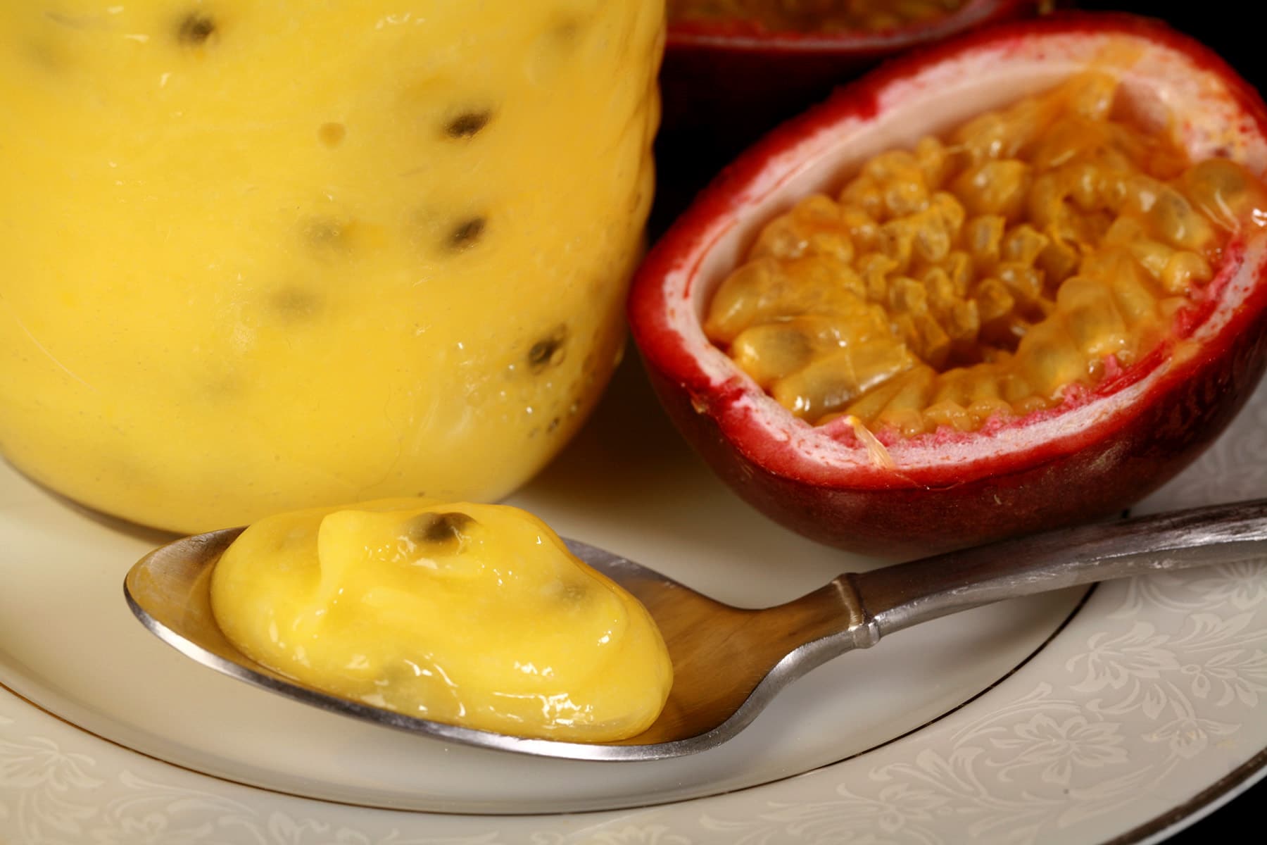 A jar of passionfruit curd on a plate, along with a spoon of curd and a sliced passionfruit.