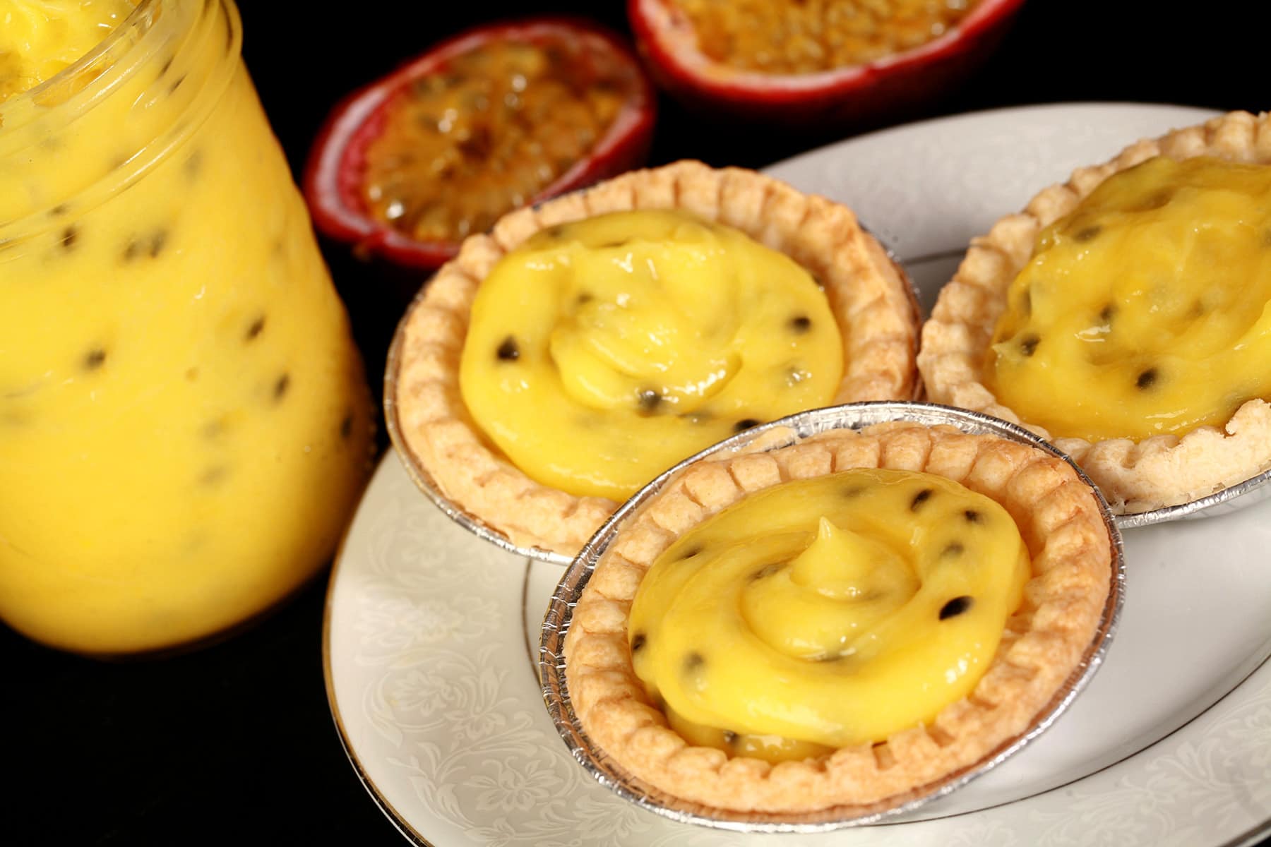 3 passionfruit tarts on a plate. There is a jar of passionfruit curd next to the plate, along with a halved passionfruit.