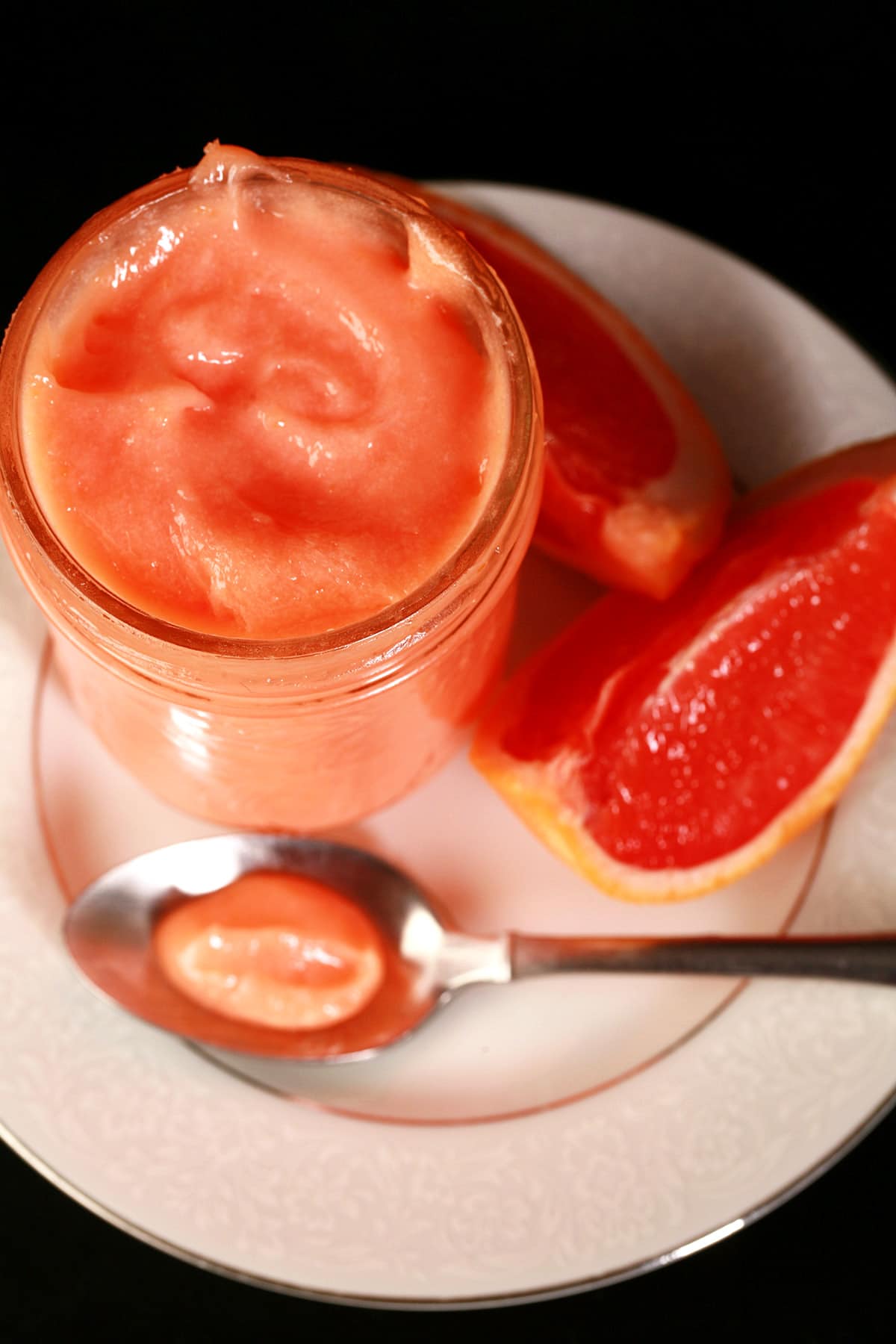 A jar of pink grapefruit curd on a plate, next to slices of pink grapefruit and a spoon of curd.