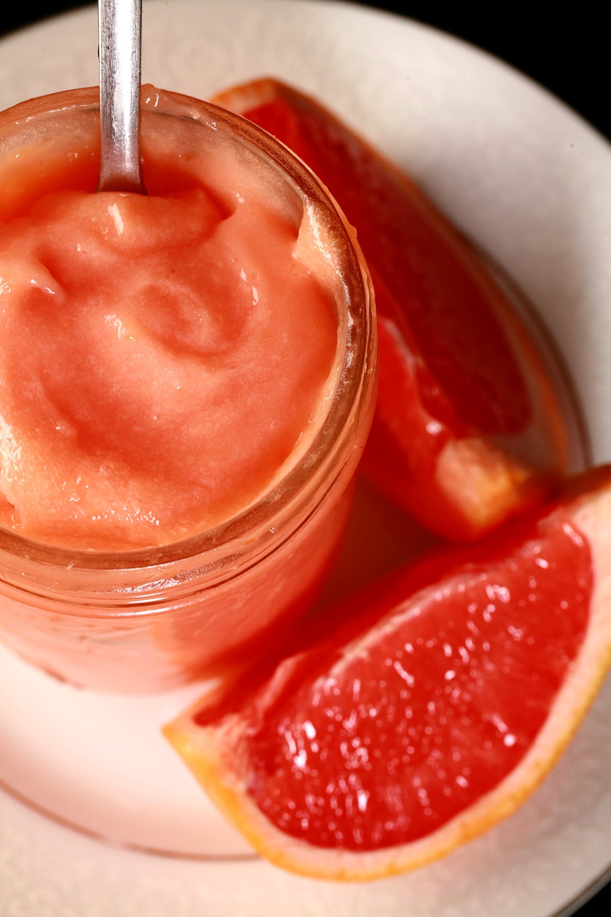 A jar of pink grapefruit curd on a plate, next to slices of pink grapefruit.
