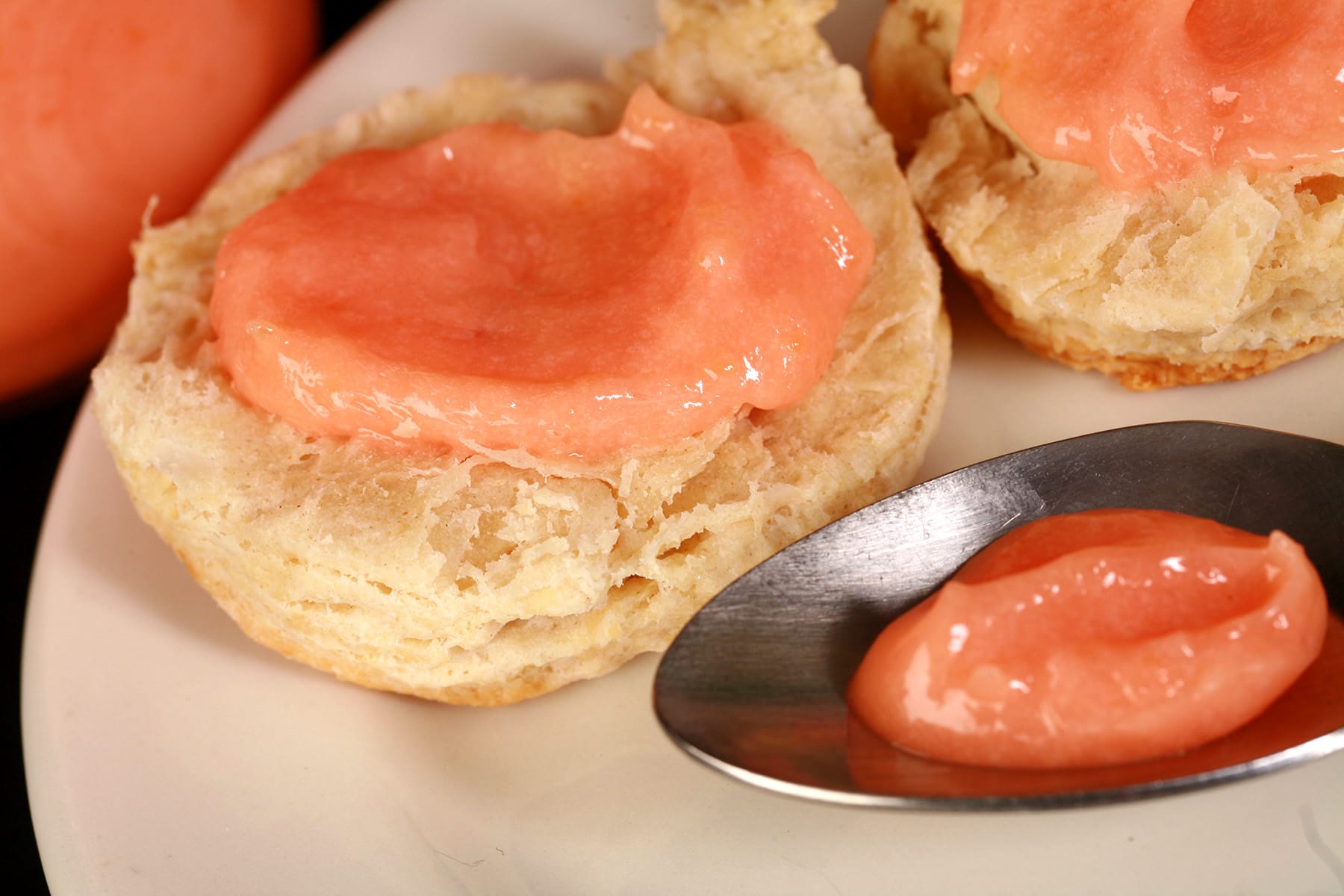 Two biscuits topped with pink grapefruit curd on a plate. There is a spoon of curd on the plate, and a jar of curd next to the plate.