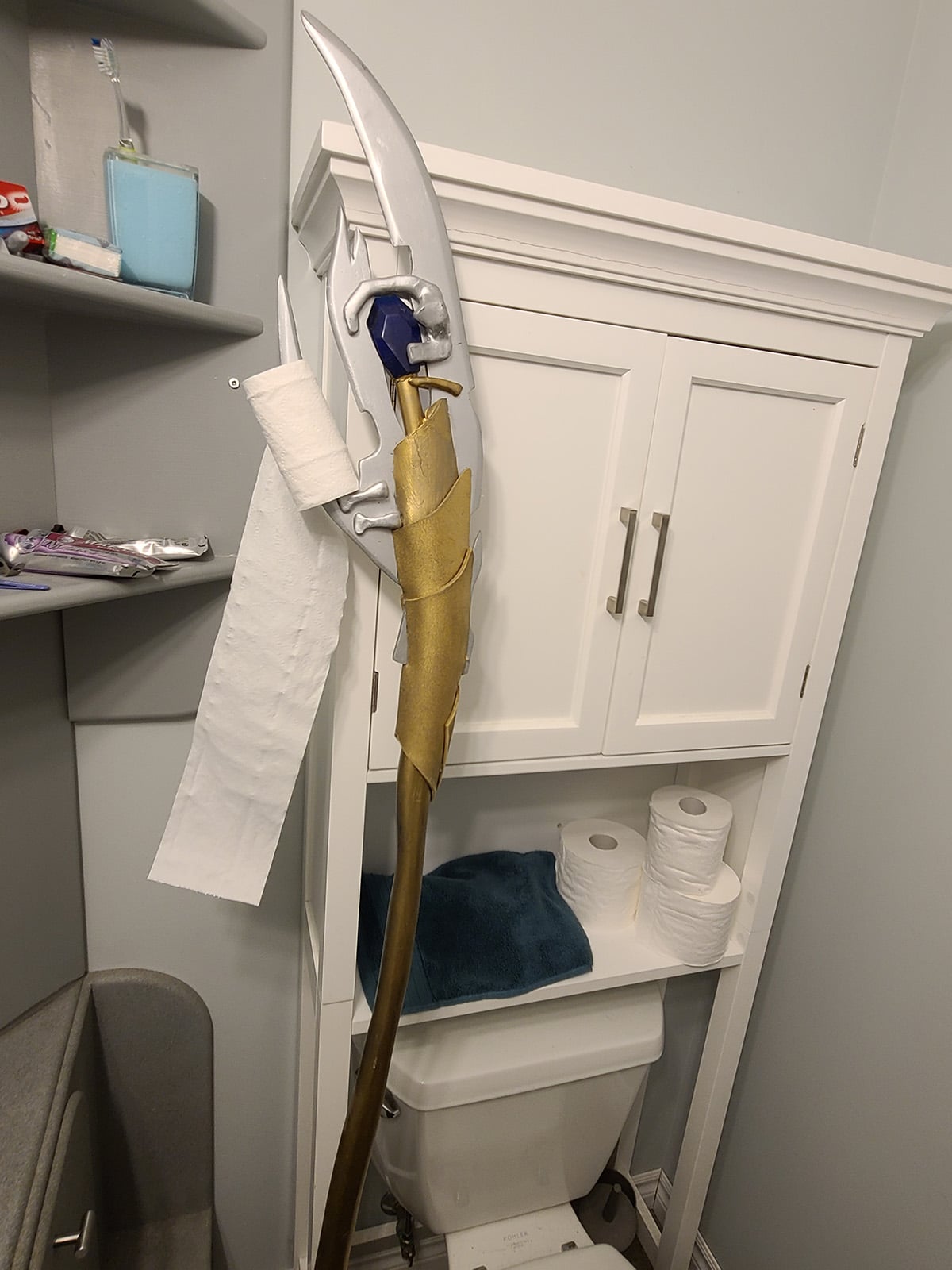 Loki's Scepter, holding a roll of toilet paper, leaning on the back of a toilet.