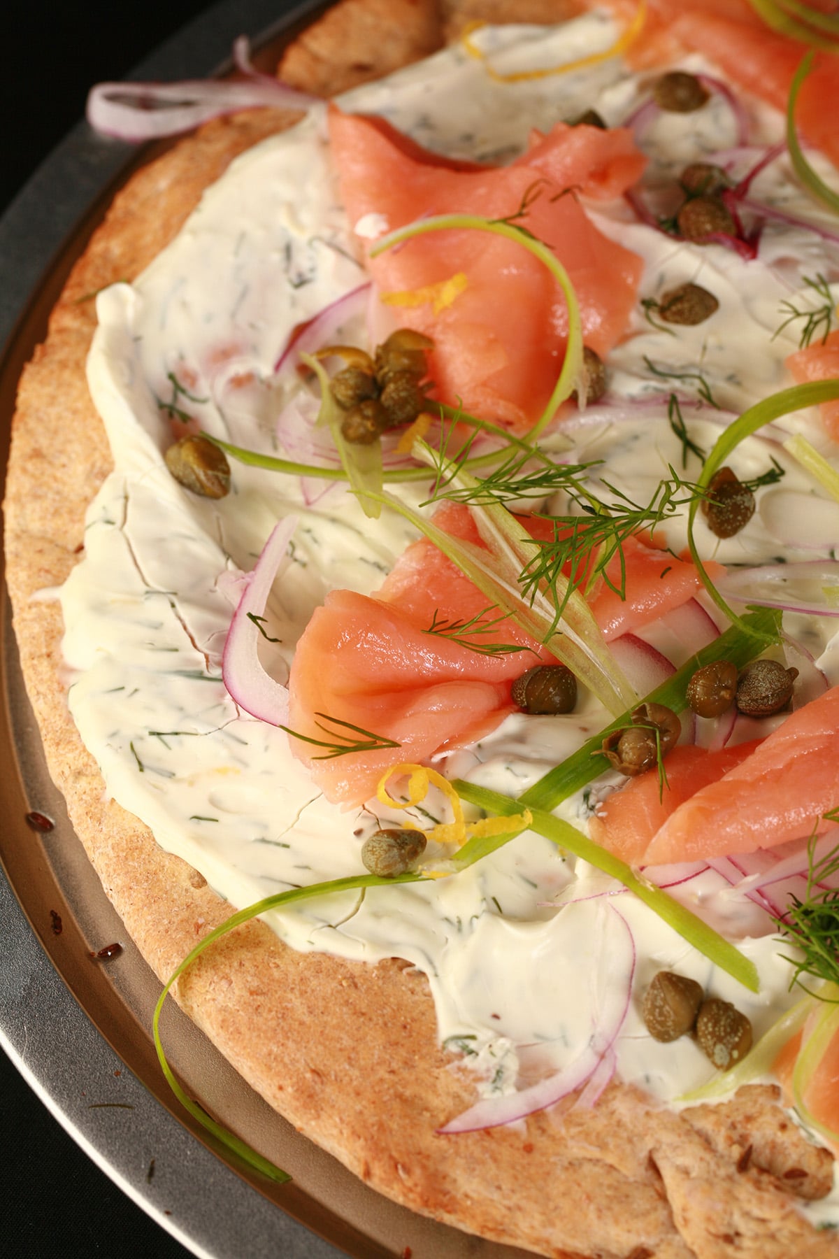 A close up view of smoked salmon pizza. The crust is rye, and it's topped with cream cheese sauce, smoked salmon, and other toppings.