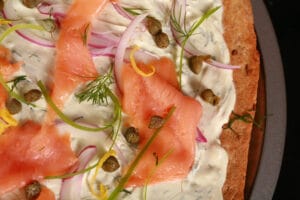 A close up view of smoked salmon pizza. The crust is rye, and it's topped with cream cheese sauce, smoked salmon, and other toppings.