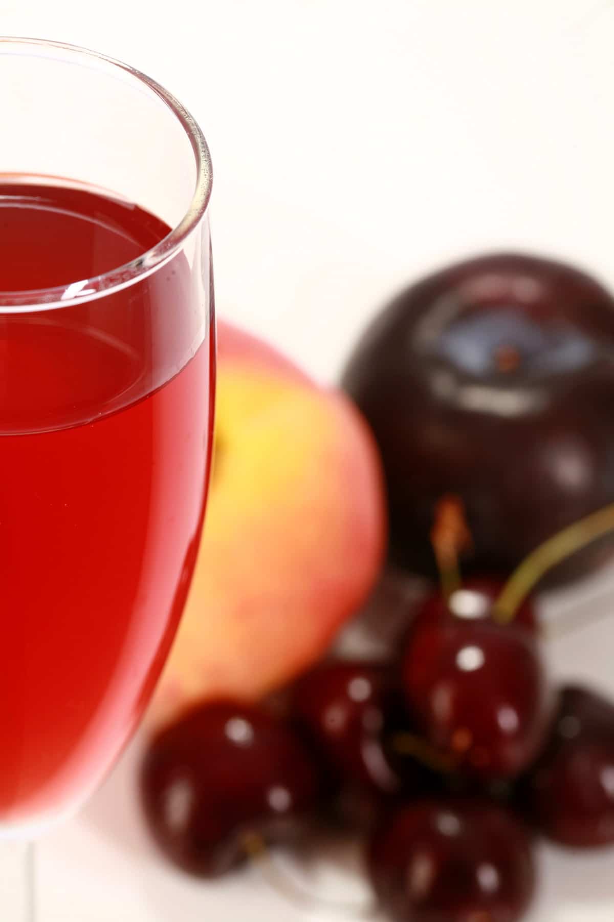 A tall glass of fruit punch-coloured stone fruit wine, next to a peach, a plum, and a few cherries.