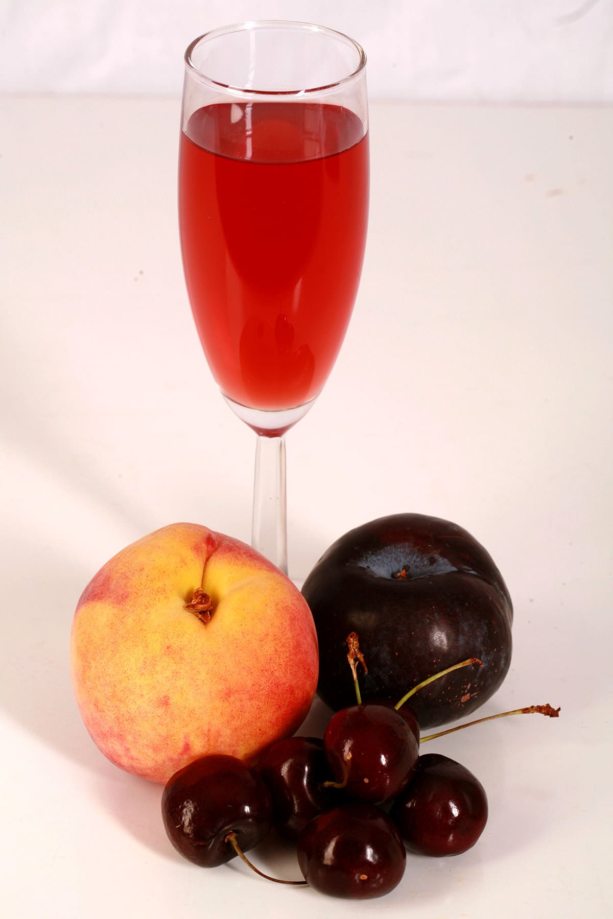 A tall glass of fruit punch-coloured stone fruit wine, next to a peach, a plum, and a few cherries.