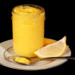 A jam jar of lemon curd on a plate, along with lemon slices and a spoon of curd.