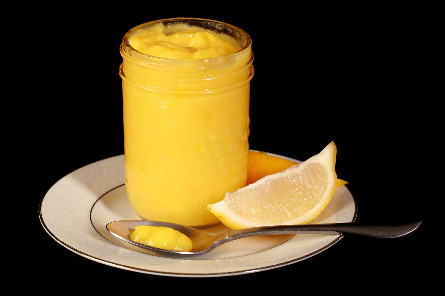 A jam jar of lemon curd on a plate, along with lemon slices and a spoon of curd.