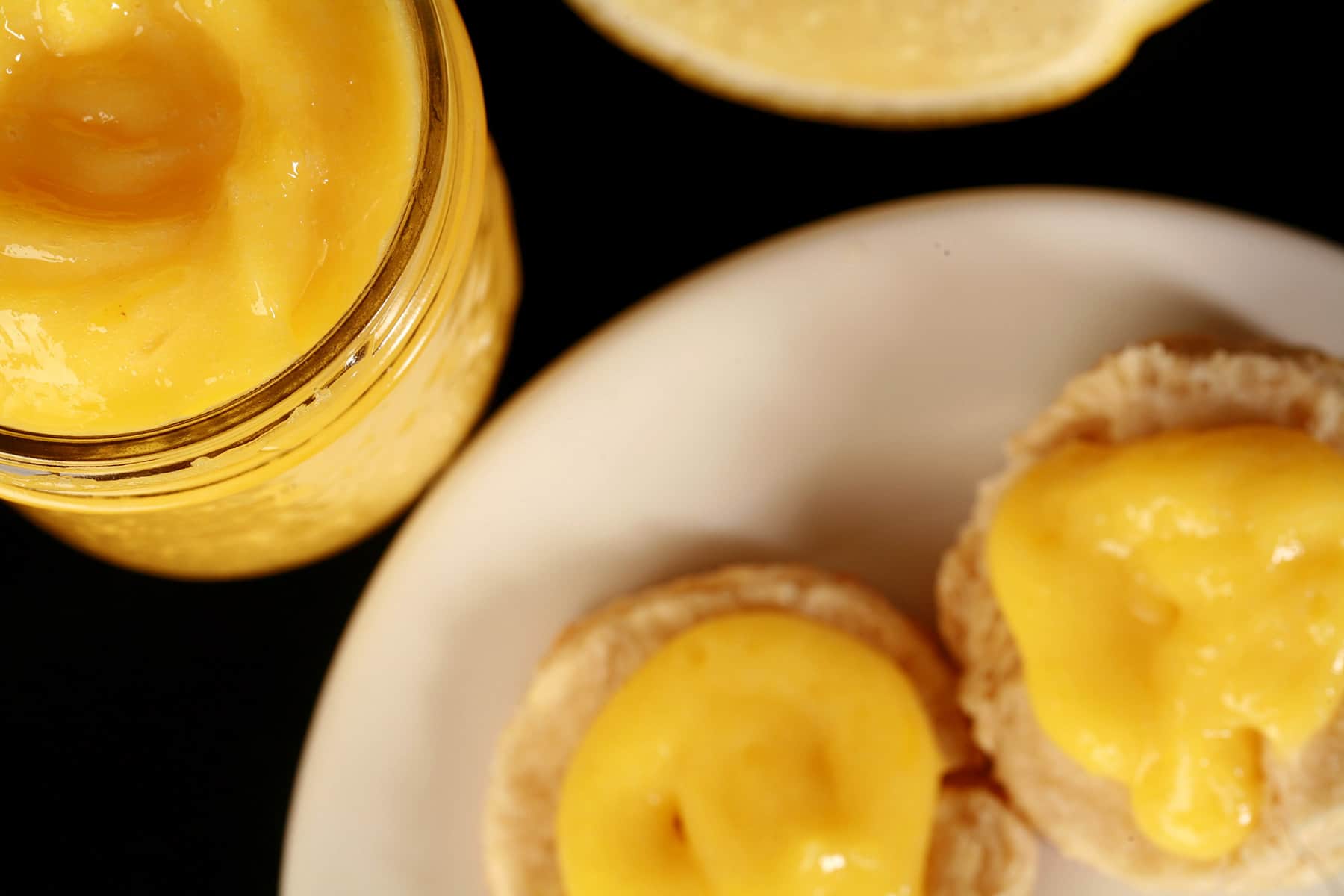 A jam jar of lemon curd on a plate, along with 2 biscuits topped with curd.