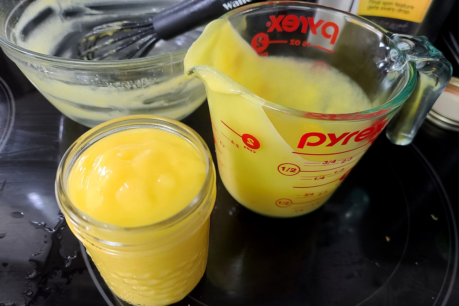 An empty mixing bowl, a glass measuring cup, and a jam jar filled with fresh lemon curd.