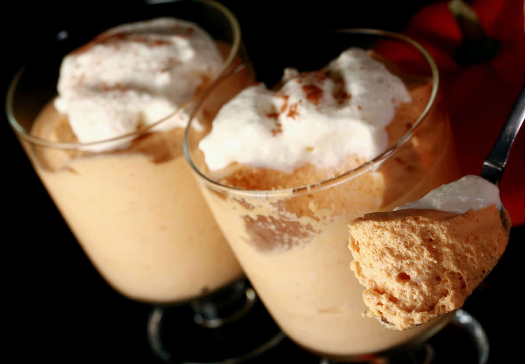 Two glasses with traditional maple pumpkin mousse in them. They are topped with whipped cream and a sprinkle of cinnamon. There is a spoon in the foreground with a generous helping of fluffy mousse.