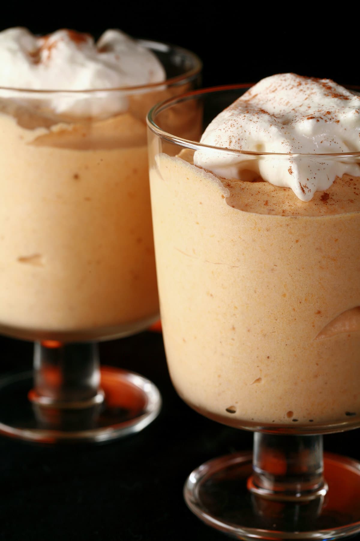 Two glasses with traditional pumpkin mousse in them. They are topped with whipped cream and a sprinkle of cinnamon.