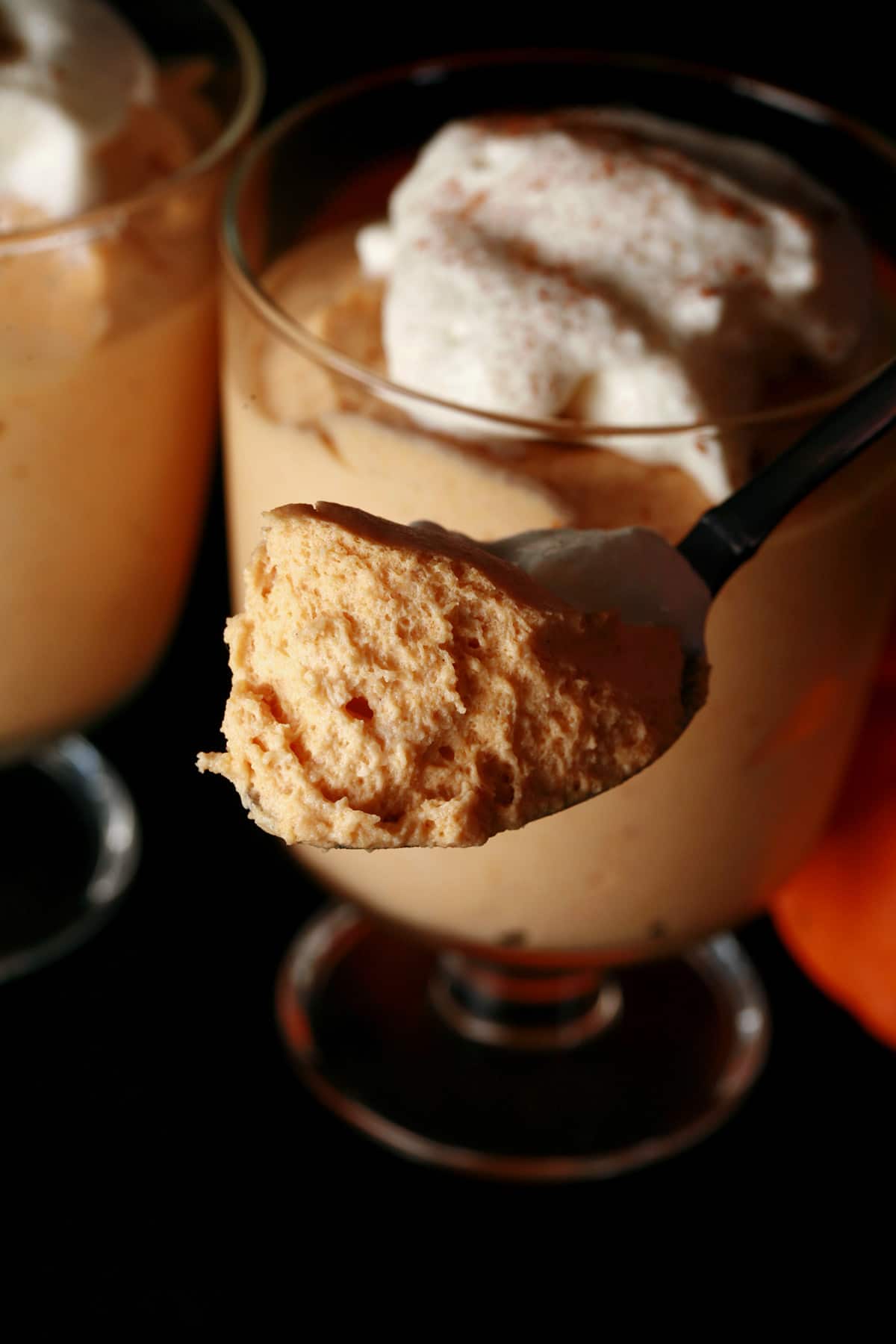 Two glasses with traditional maple pumpkin mousse in them. They are topped with whipped cream and a sprinkle of cinnamon. There is a spoon in the foreground with a generous helping of fluffy mousse.