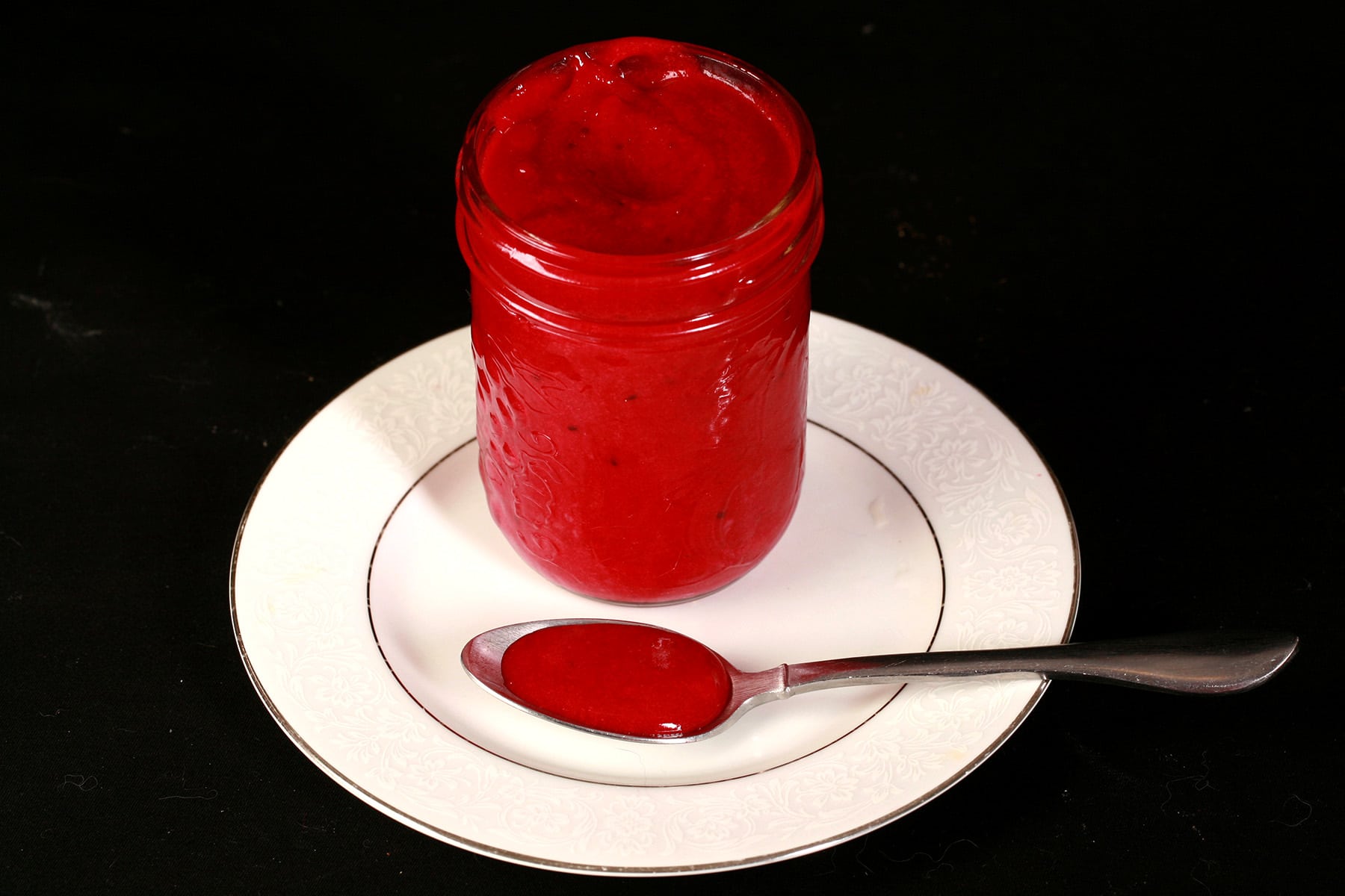 A jar of blackcurrant curd on a plate, along with a spoon of the curd.
