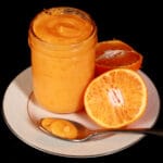 A jar of orange curd on a plate, along with a spoon of curd and a sliced orange.