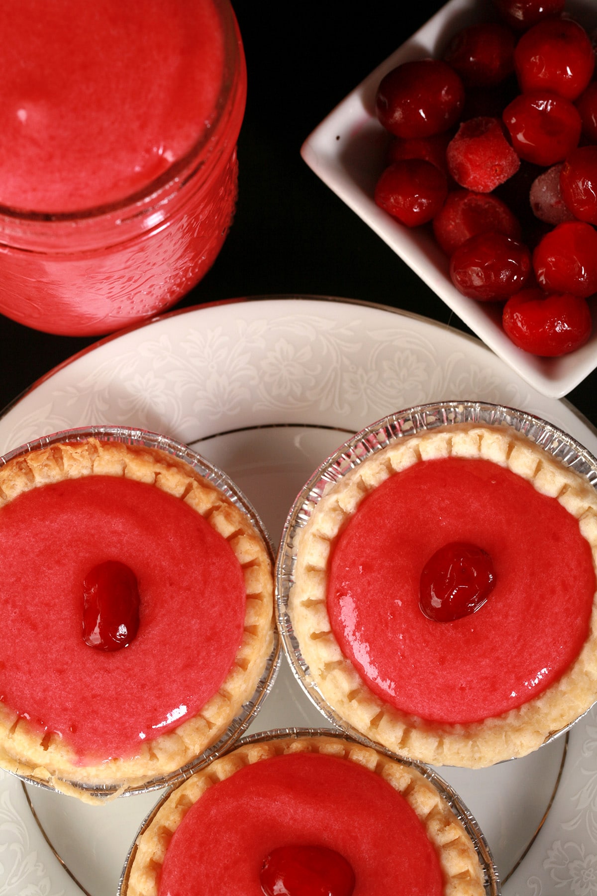 3 cranberry tarts on a plate, next to a jar of cranberry curd and a bowl of fresh cranberries.