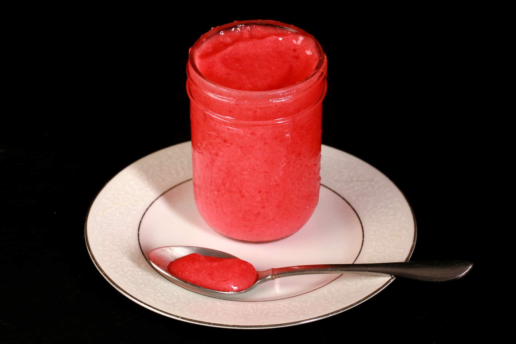 A jar of cranberry curd on a small white plate, next to a spoon with curd on it.