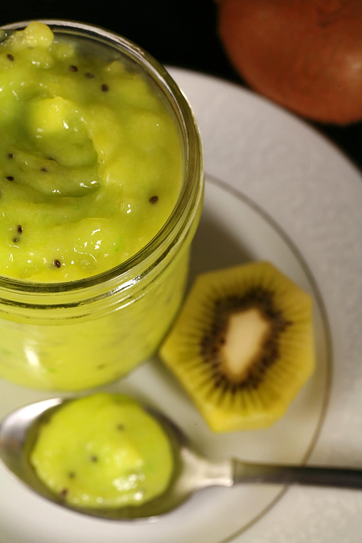 A jar of kiwi curd on a plate. There is a spoon of curd next to the jar.