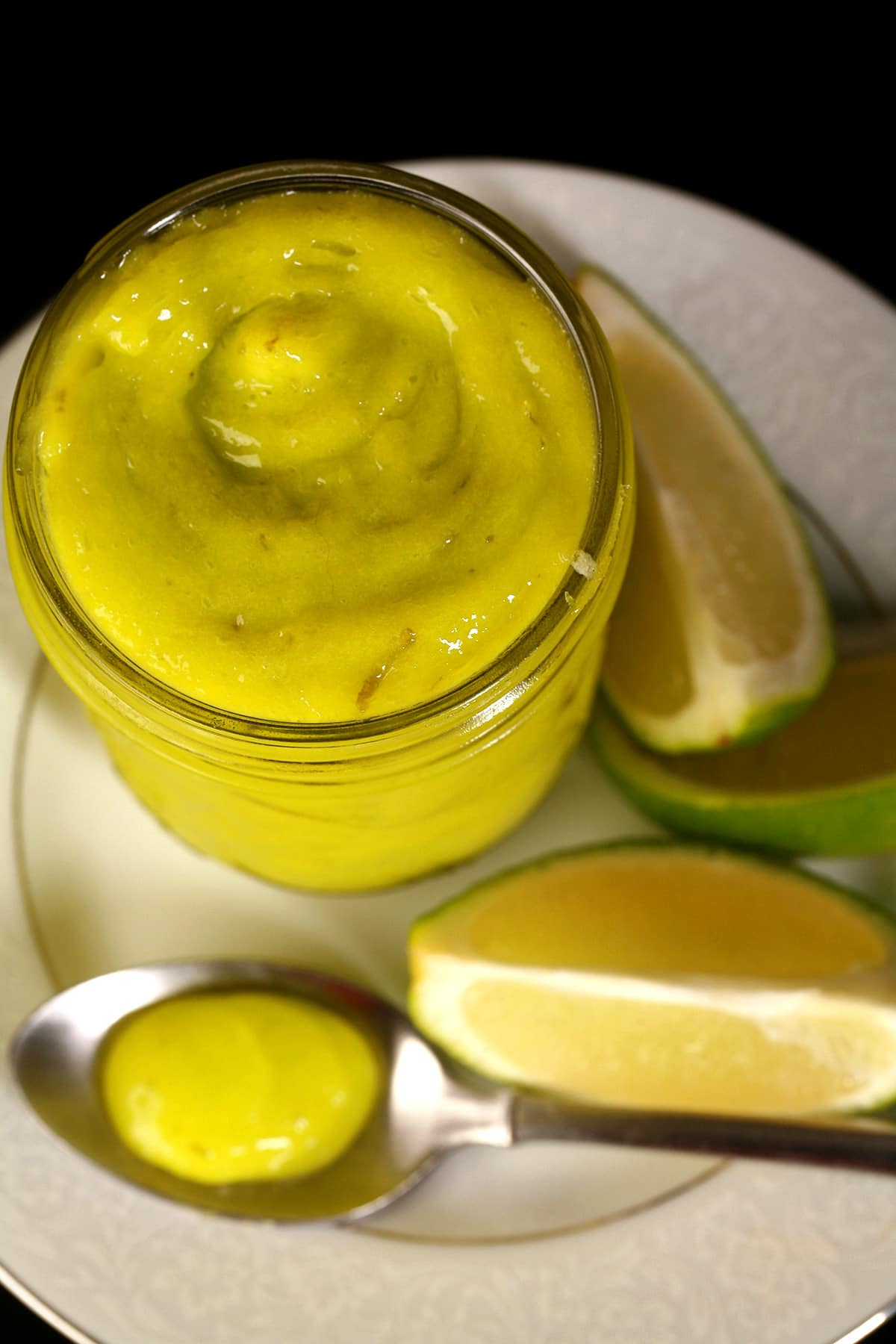 A jam jar of lime curd on a plate with fresh lime slices.