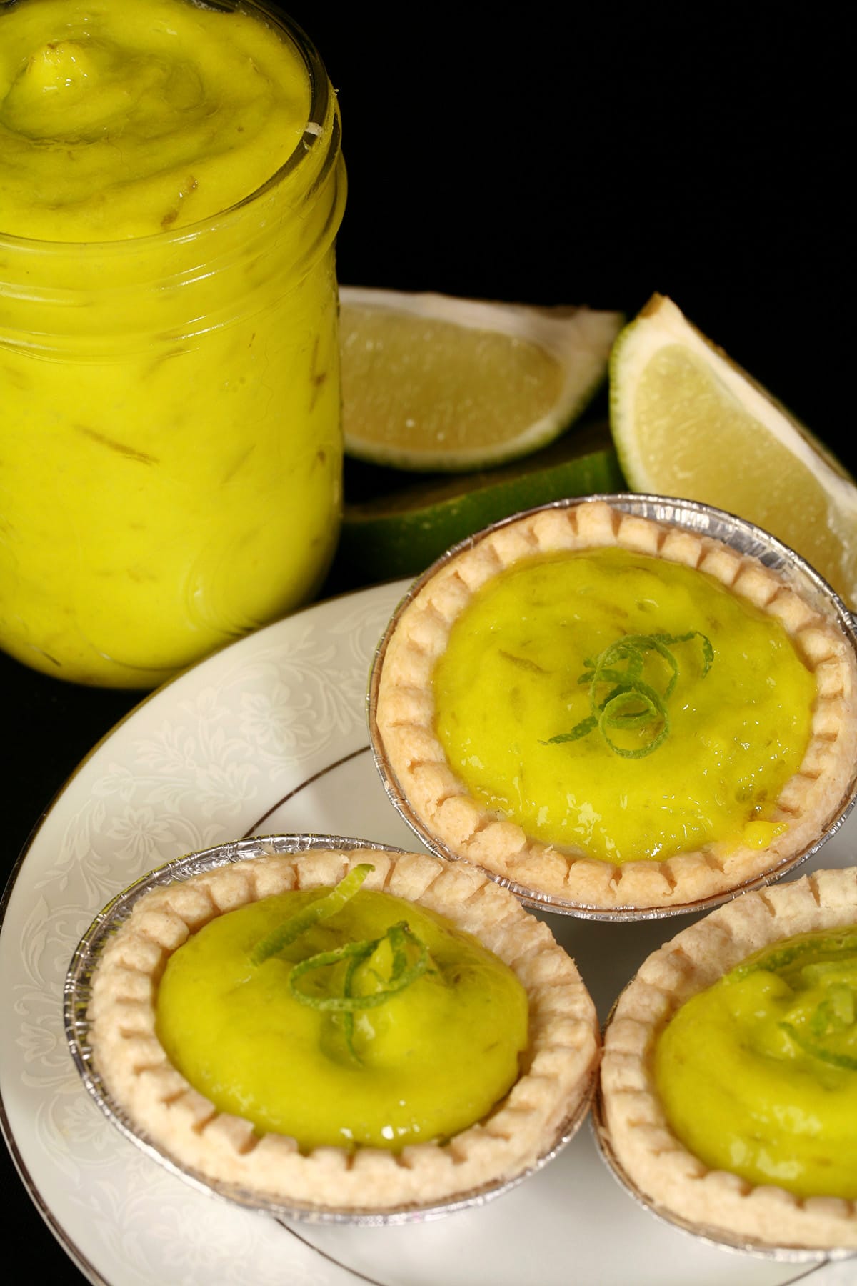 A glass jam jar of lime curd next to a plate of mini lime tarts.