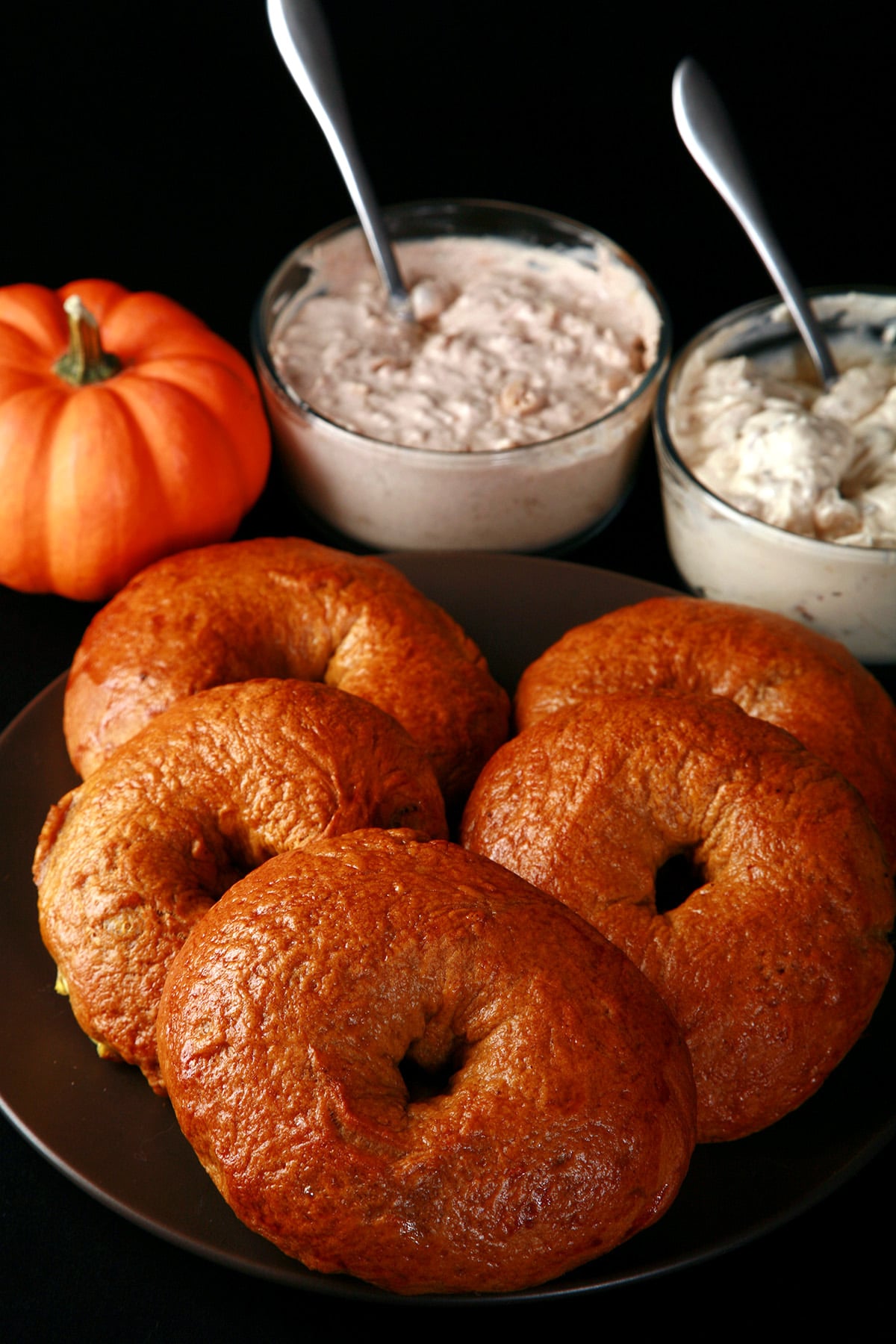 A plate of maple pumpkin spiced bagels. There is a mini pumpkin and 2 dishes of flavoured cream cheese behind the plate.