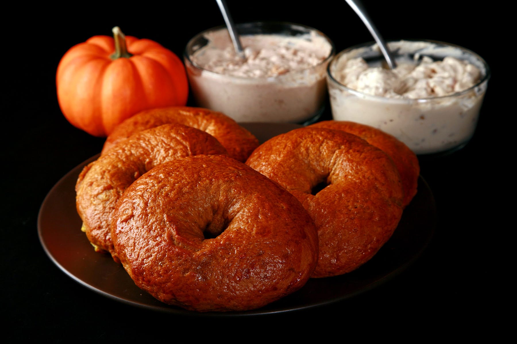 A plate of maple pumpkin spiced bagels. There is a mini pumpkin and 2 dishes of flavoured cream cheese behind the plate.