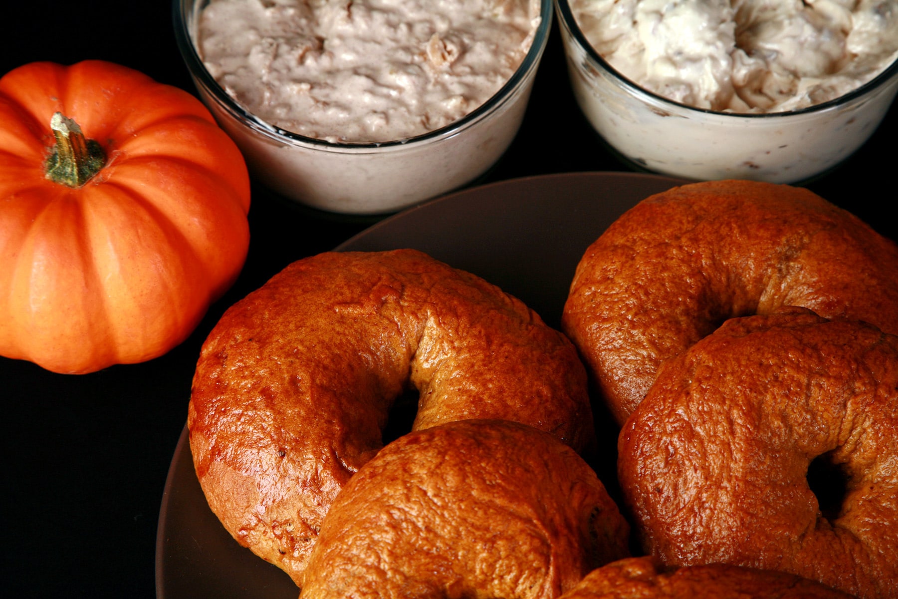 A plate of maple pumpkin spiced bagels. There is a mini pumpkin and 2 dishes of flavoured cream cheese behind the plate.