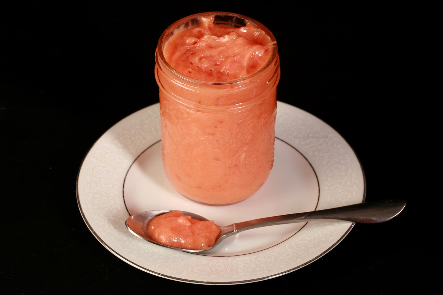 A jam jar of raspberry curd on a plate, next to a spoon with the pink curd on it.