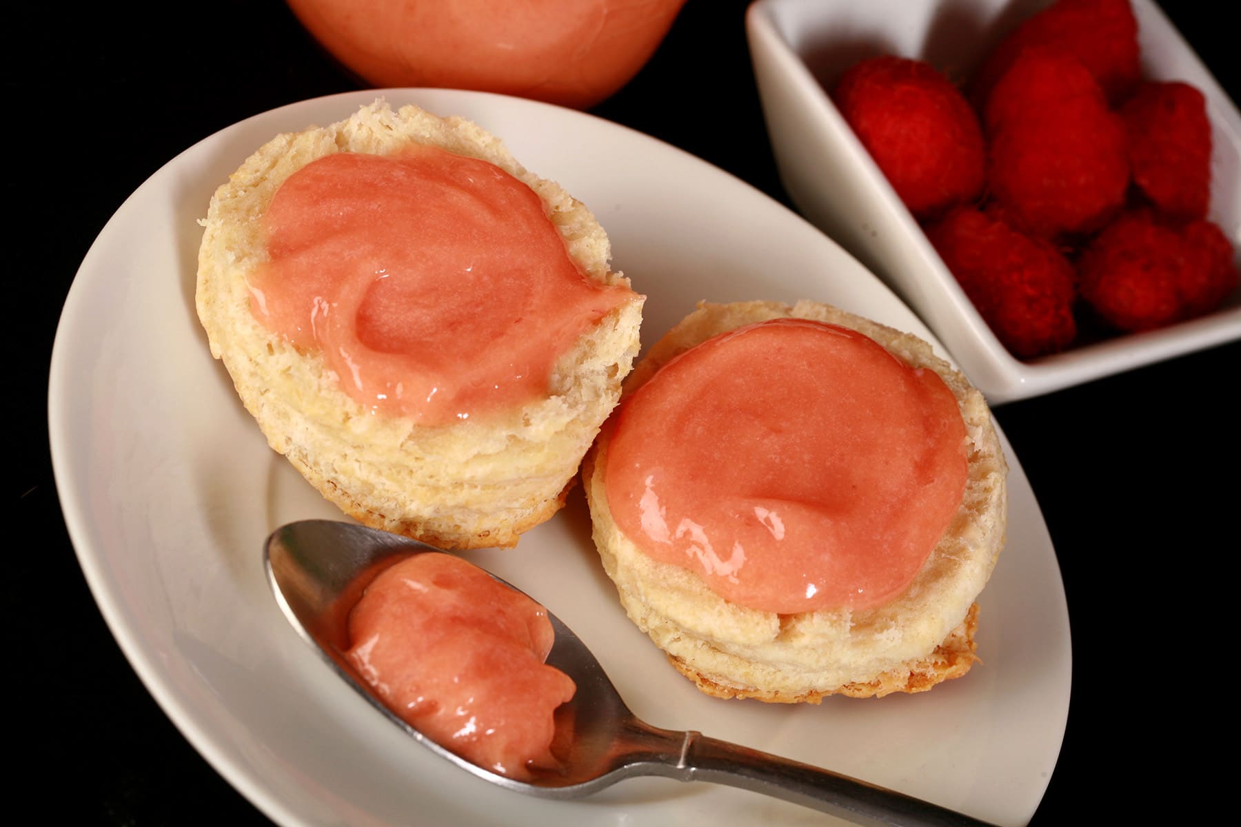 Two raspberry curd covered biscuits on a plate, along with a spoon of the pink curd.