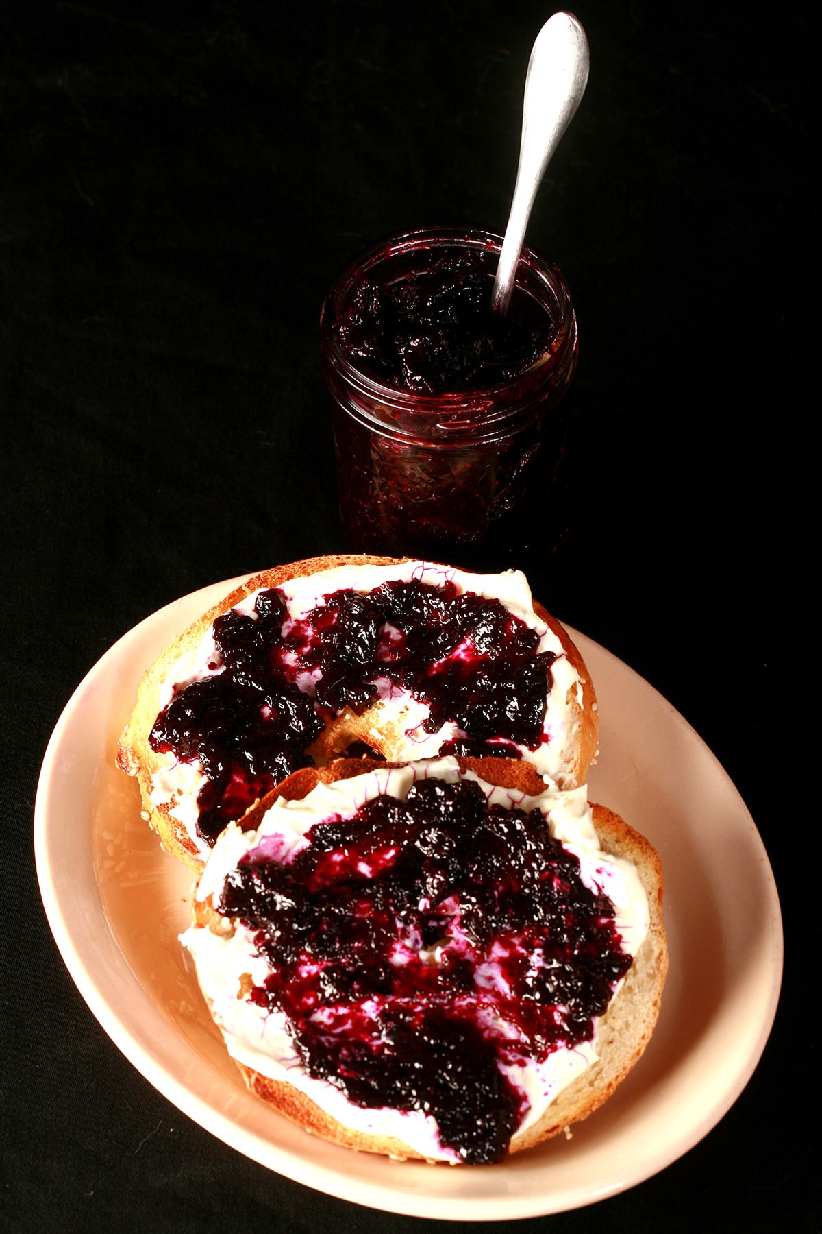 A bagel spread with cream cheese and topped with this small batch blueberry jam. There is a small jar of jam next to it.