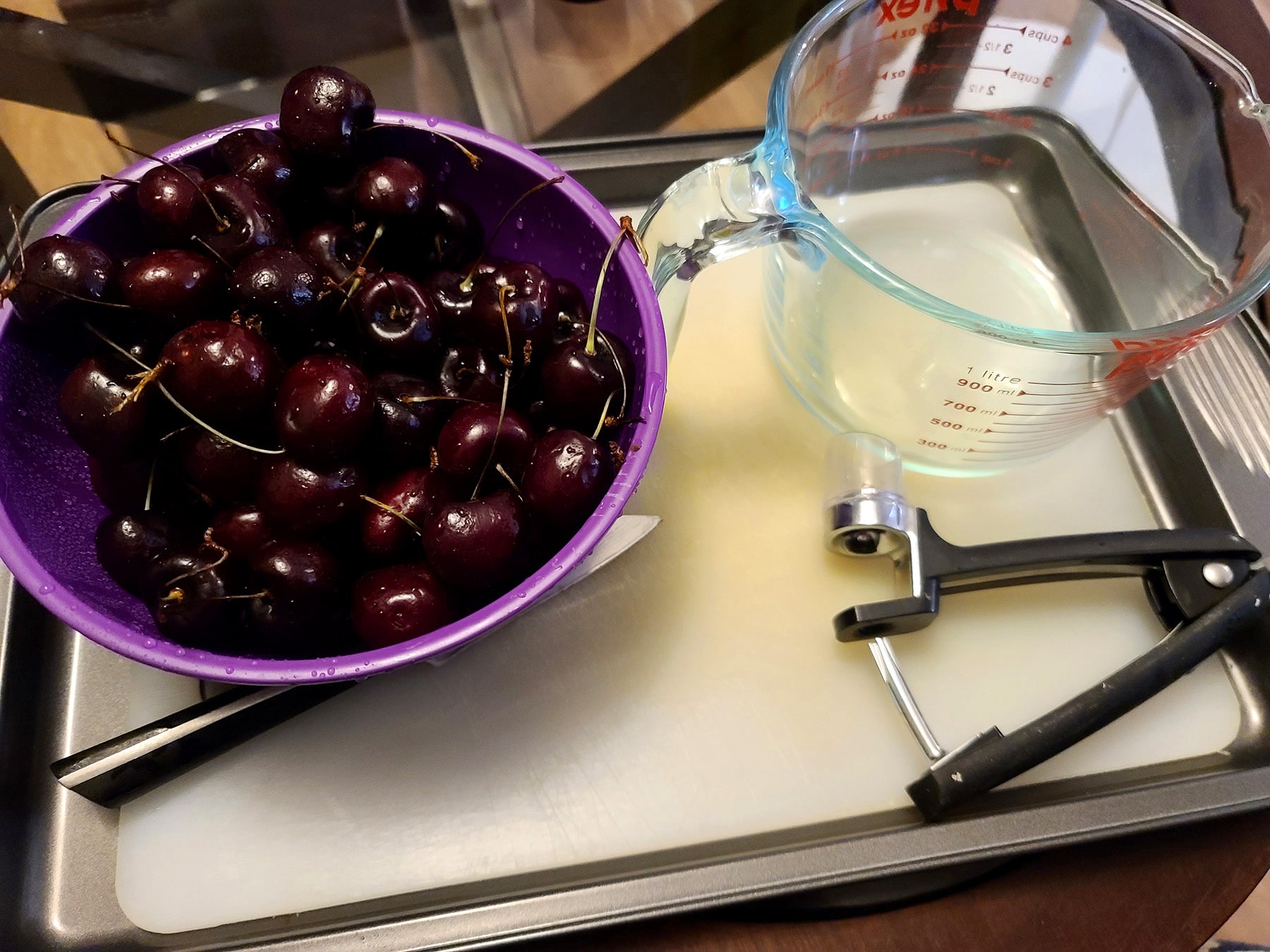 A bowl of cherries on a baking pan, along with a cherry pitter and a measuring cup.