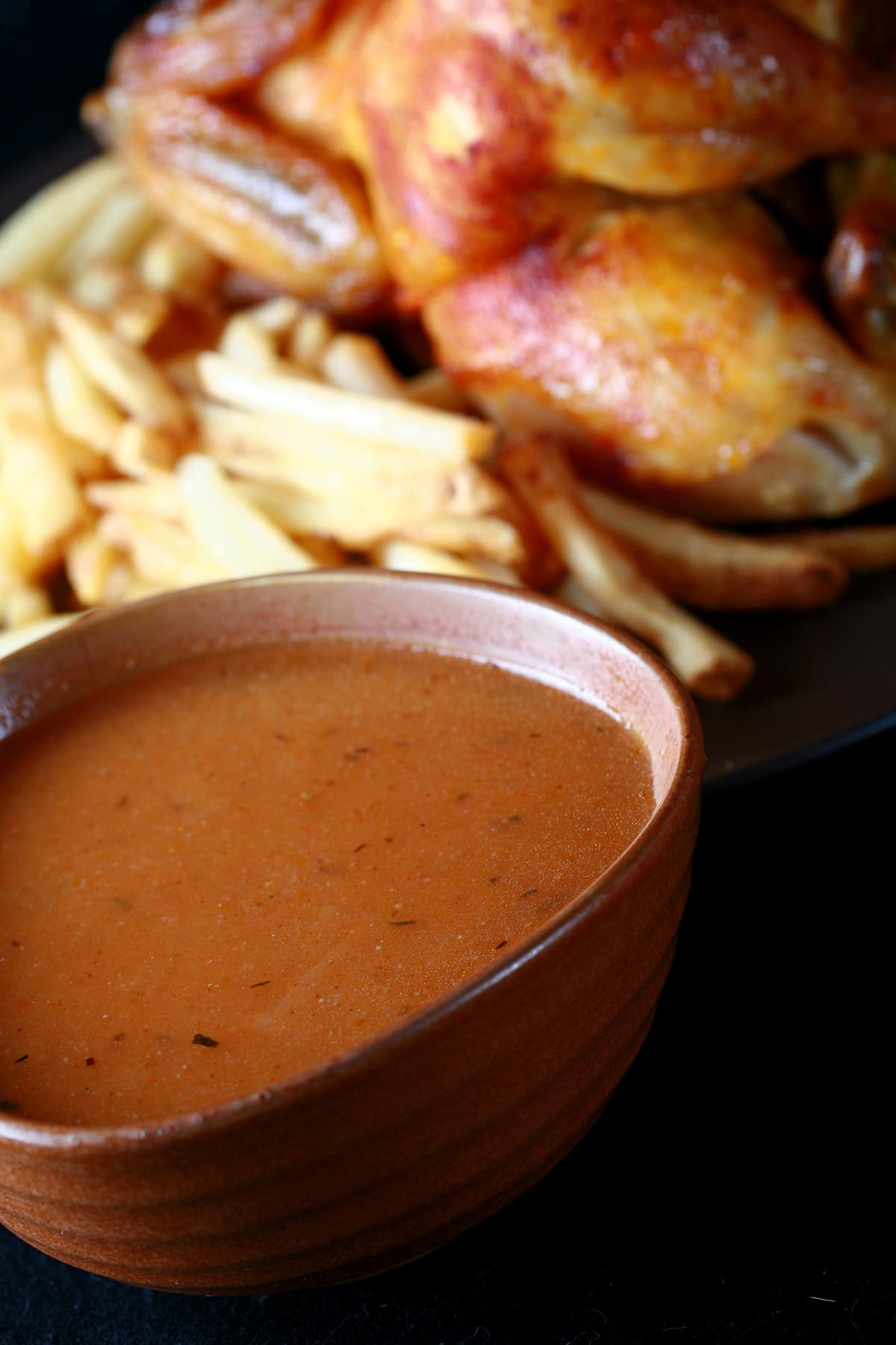 A large bowl of homemade Swiss Chalet Sauce next to a roasted chicken and fries.