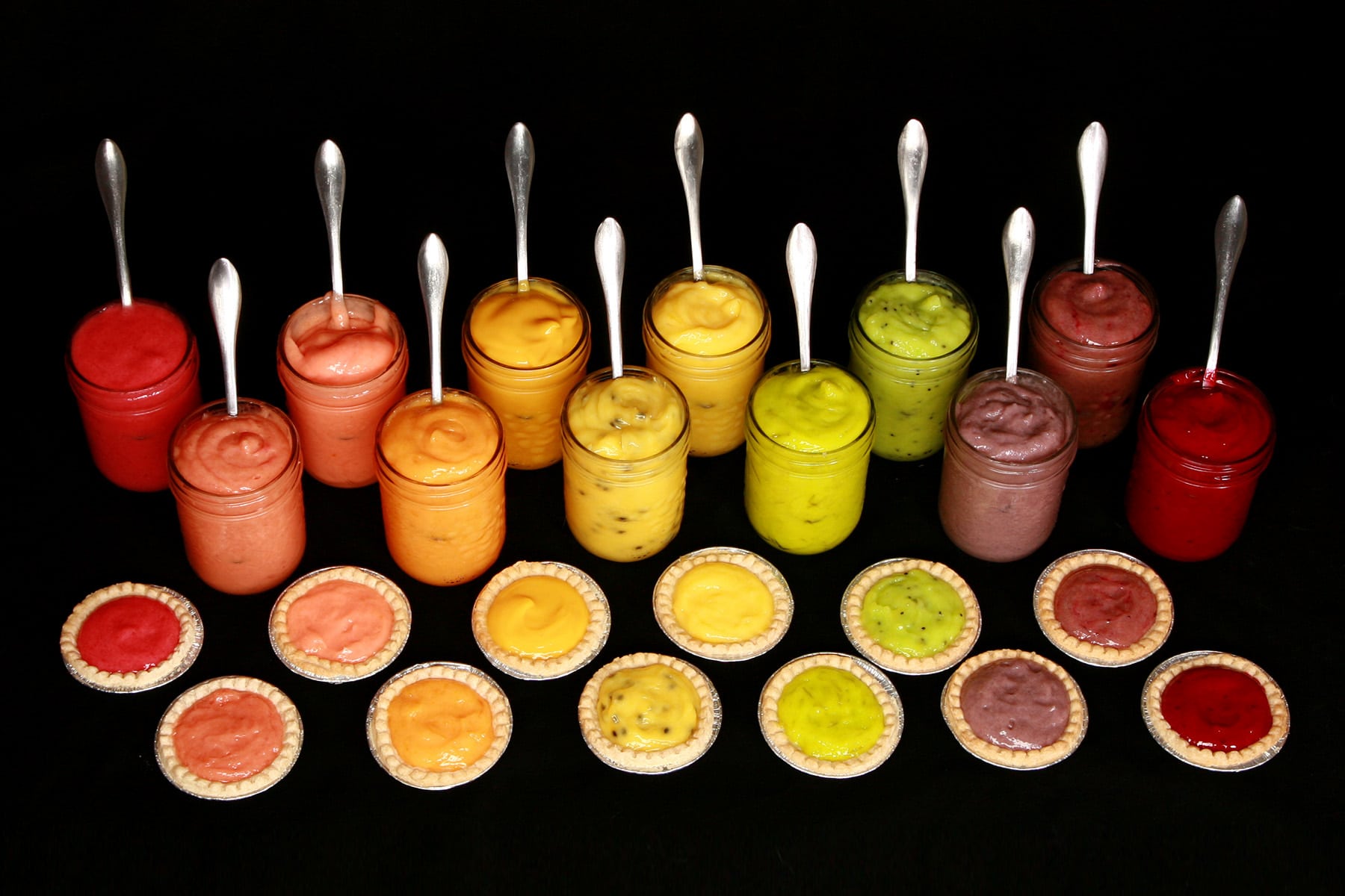 A row of open jars of fruit curds - arranged in rainbow order - each with a fruit tart in front of it.
