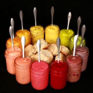 A circle of jam jars filled with fruit curds surround a plate of biscuits. The jars are open, each has a spoon in it, and they are arranged in rainbow order.